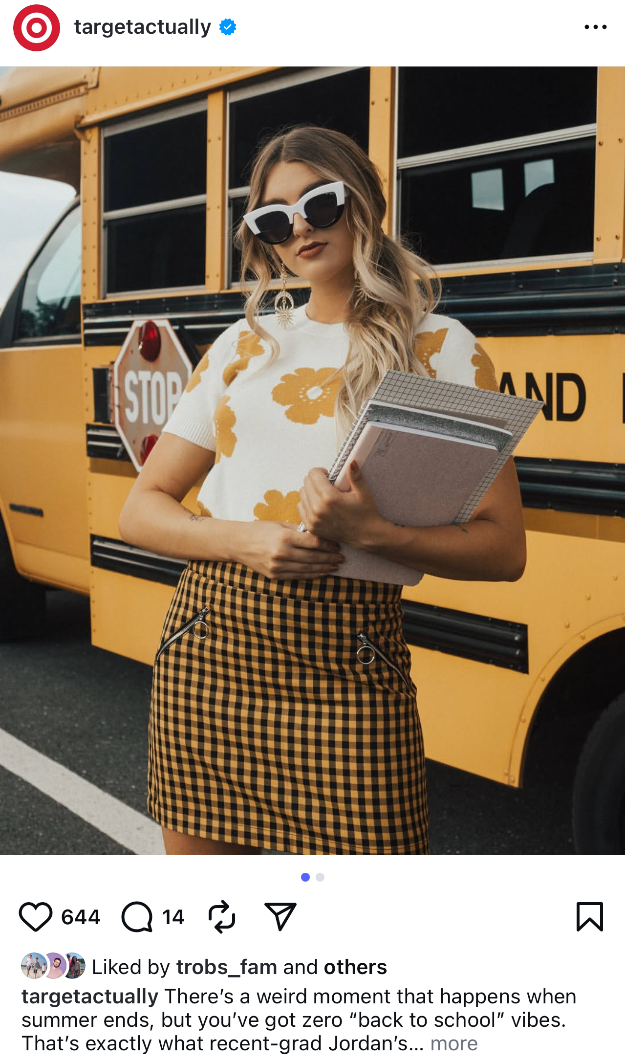 A young woman with wavy blonde hair wearing large black and white sunglasses, a white t-shirt with orange floral print, and a yellow and black gingham skirt, standing in front of a yellow school bus, holding notebooks.