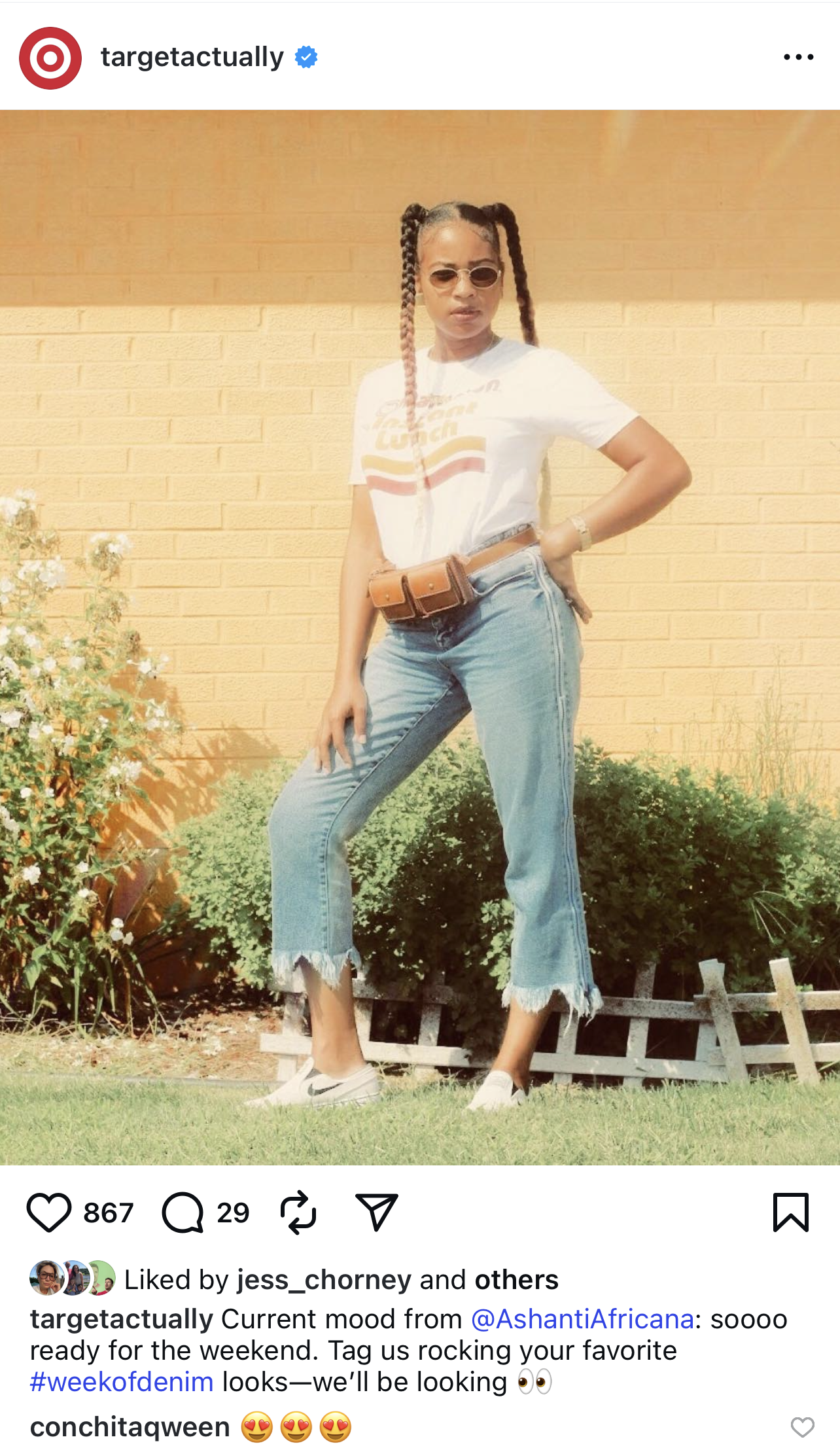 Woman with braided hair wearing sunglasses, white graphic t-shirt, high-waisted distressed jeans, and sneakers, standing outdoors in front of a brick wall, with greenery and flowers nearby.