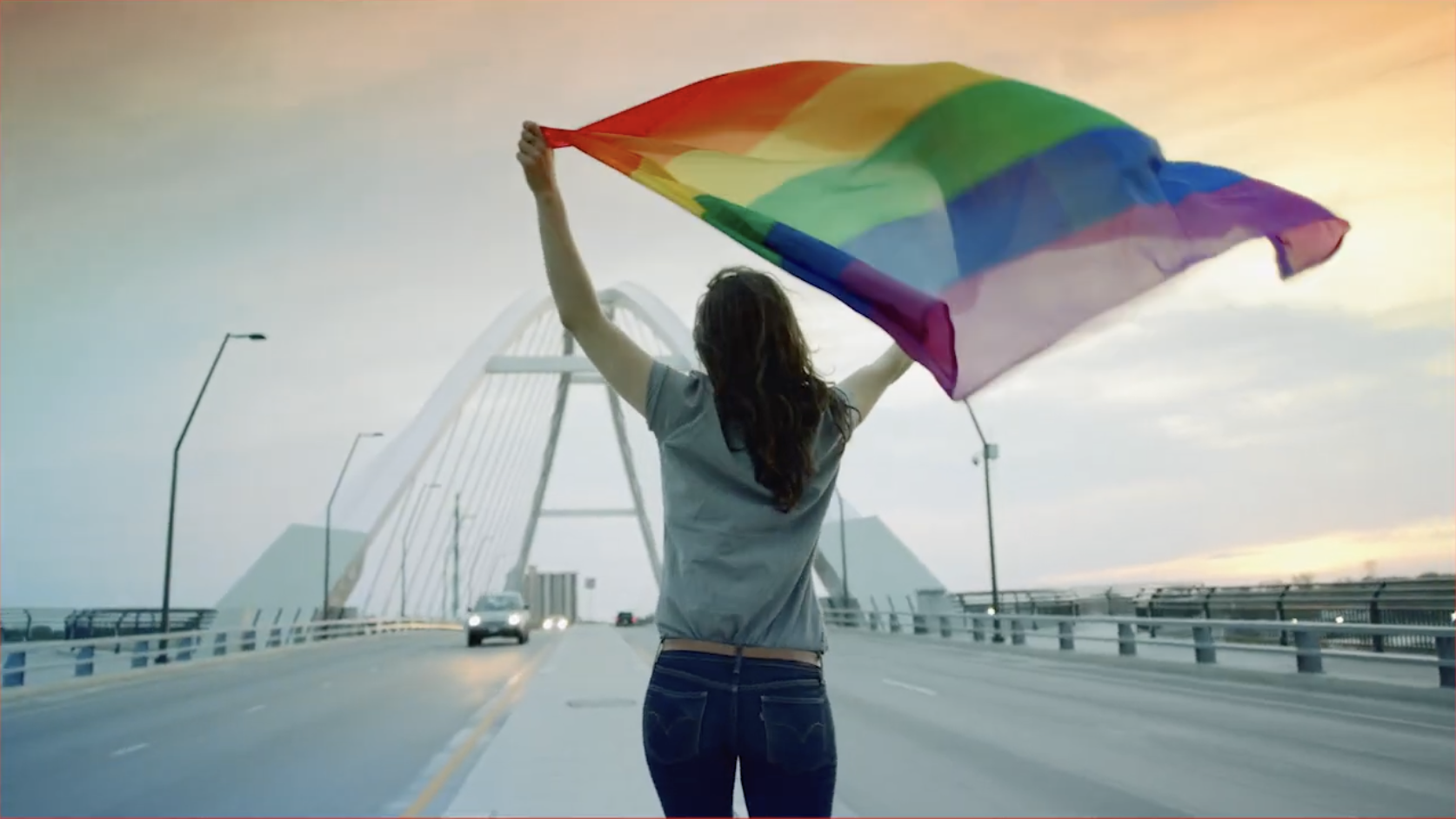 Person standing on a bridge holding a rainbow pride flag, with a modern white arched bridge structure in the background during sunset.