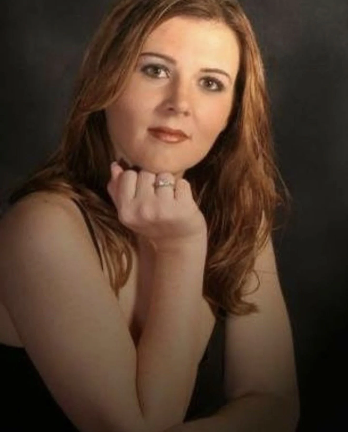 A woman with long, wavy red hair, wearing a black top, resting her chin on her hand with a silver ring, posing against a dark background.