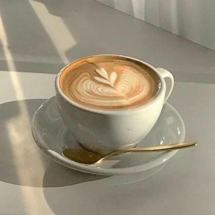 A cup of coffee with latte art on top, placed on a saucer with a golden spoon, on a white table.