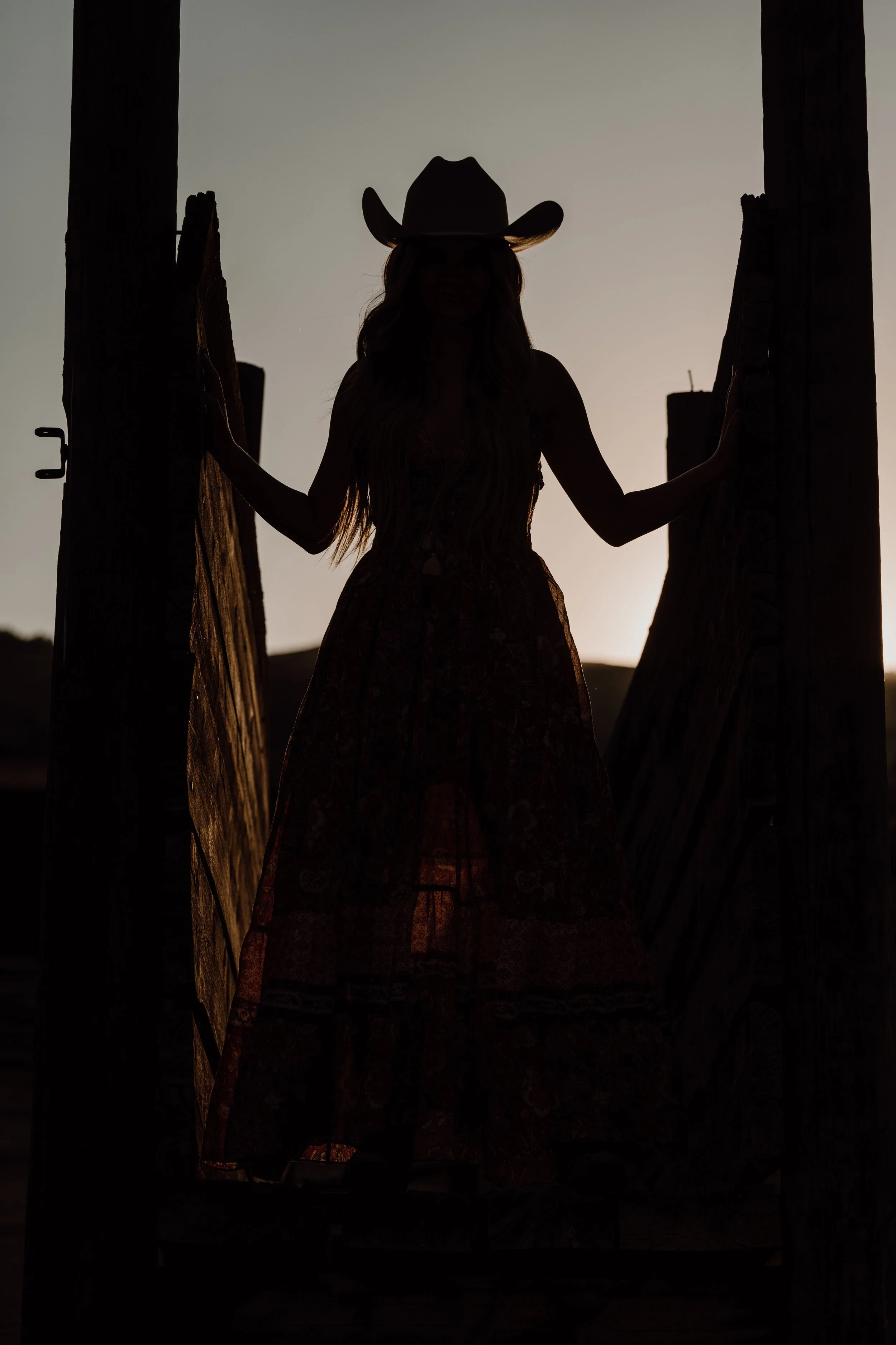 Silhouette of a woman wearing a cowboy hat, standing between two wooden posts at sunset or sunrise.