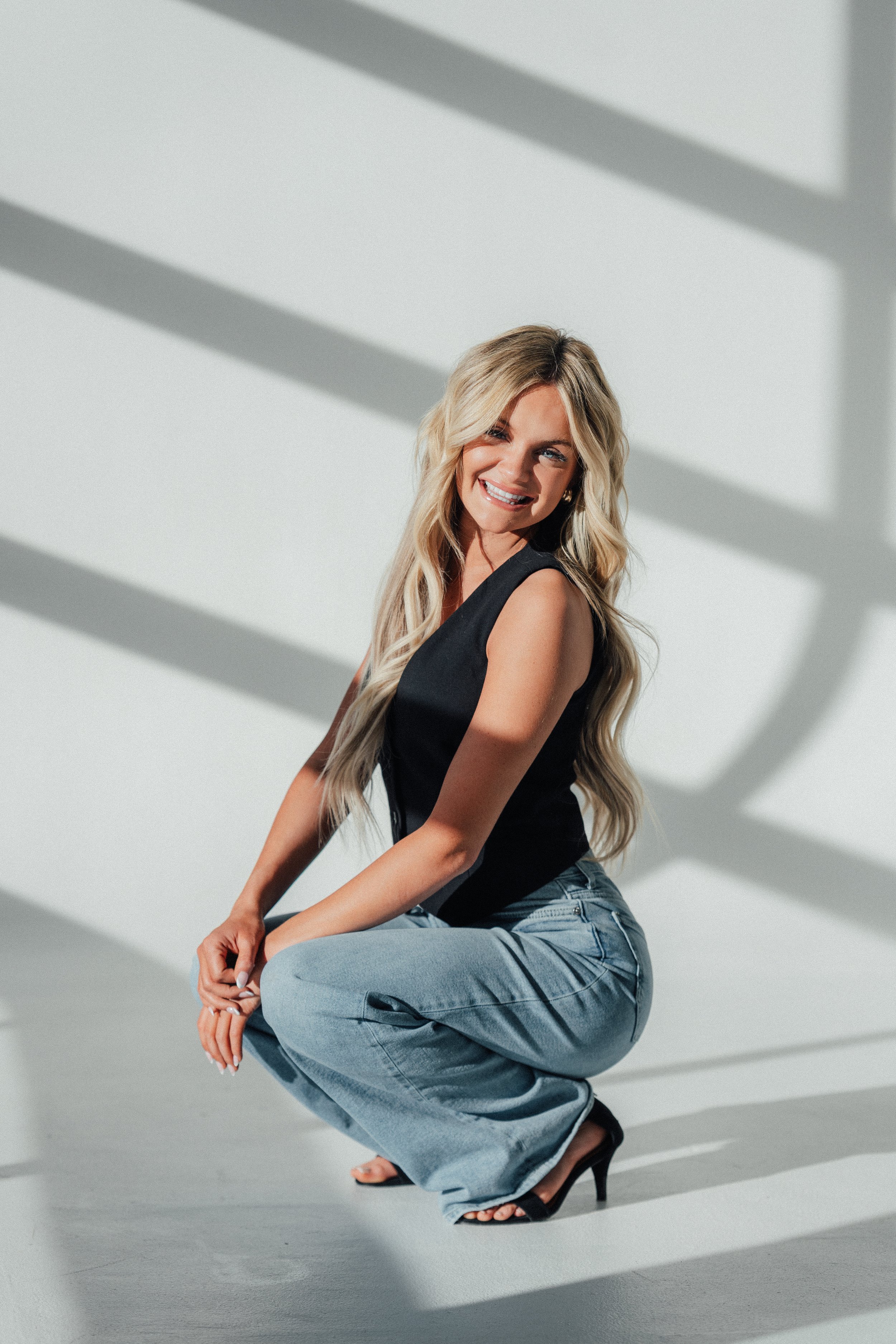 A woman in black top and blue jeans squatting and smiling, with sunlight casting shadows on a white wall.
