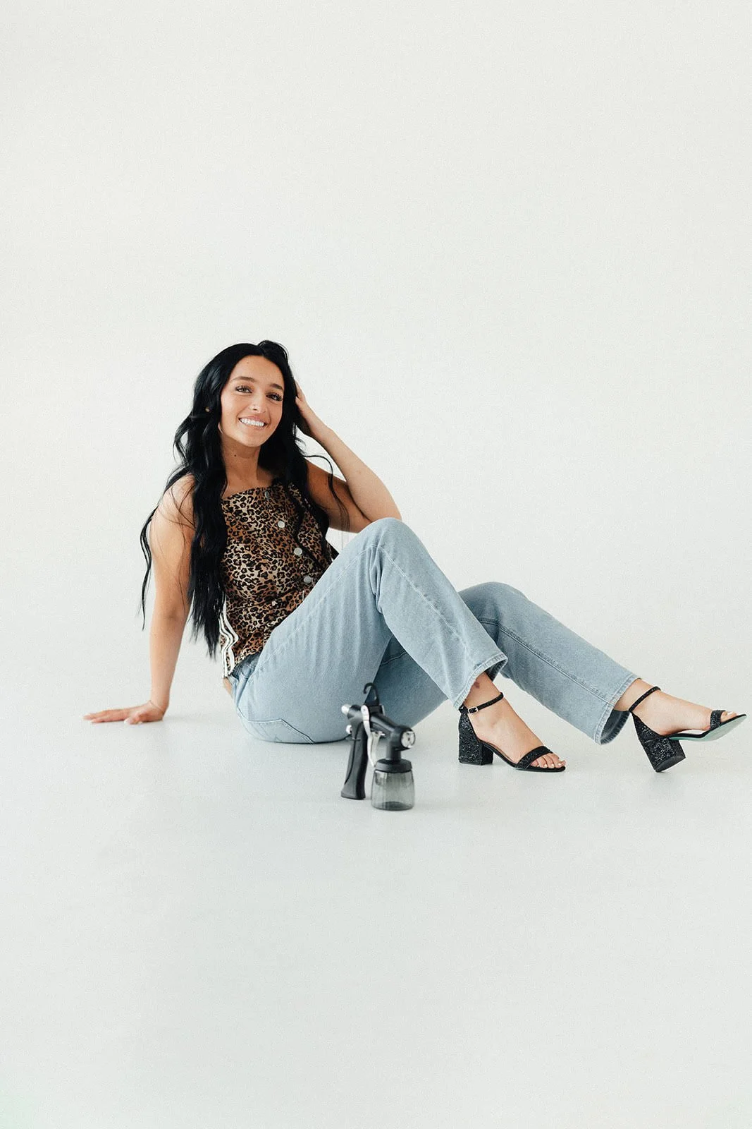 A woman with long black hair smiling, sitting on a white studio floor near a spray paint tattoo gun and a small spray bottle, dressed in a leopard print top, light wash jeans, and black high-heeled sandals.