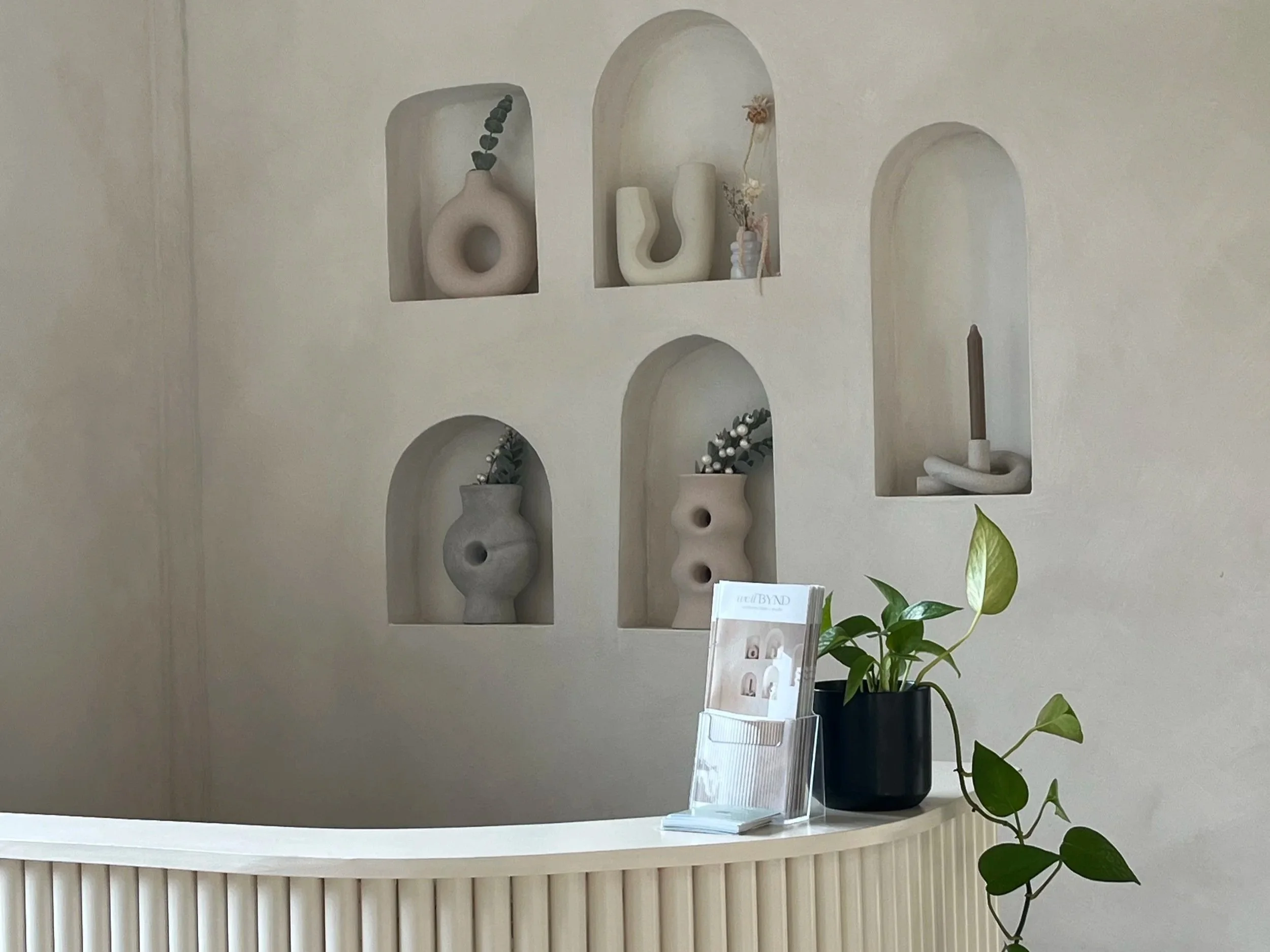A white curved display shelf with five alcoves, holding modern decorative vases and a small dried flower arrangement, against a neutral wall. There is a potted green plant on a white surface in the foreground.