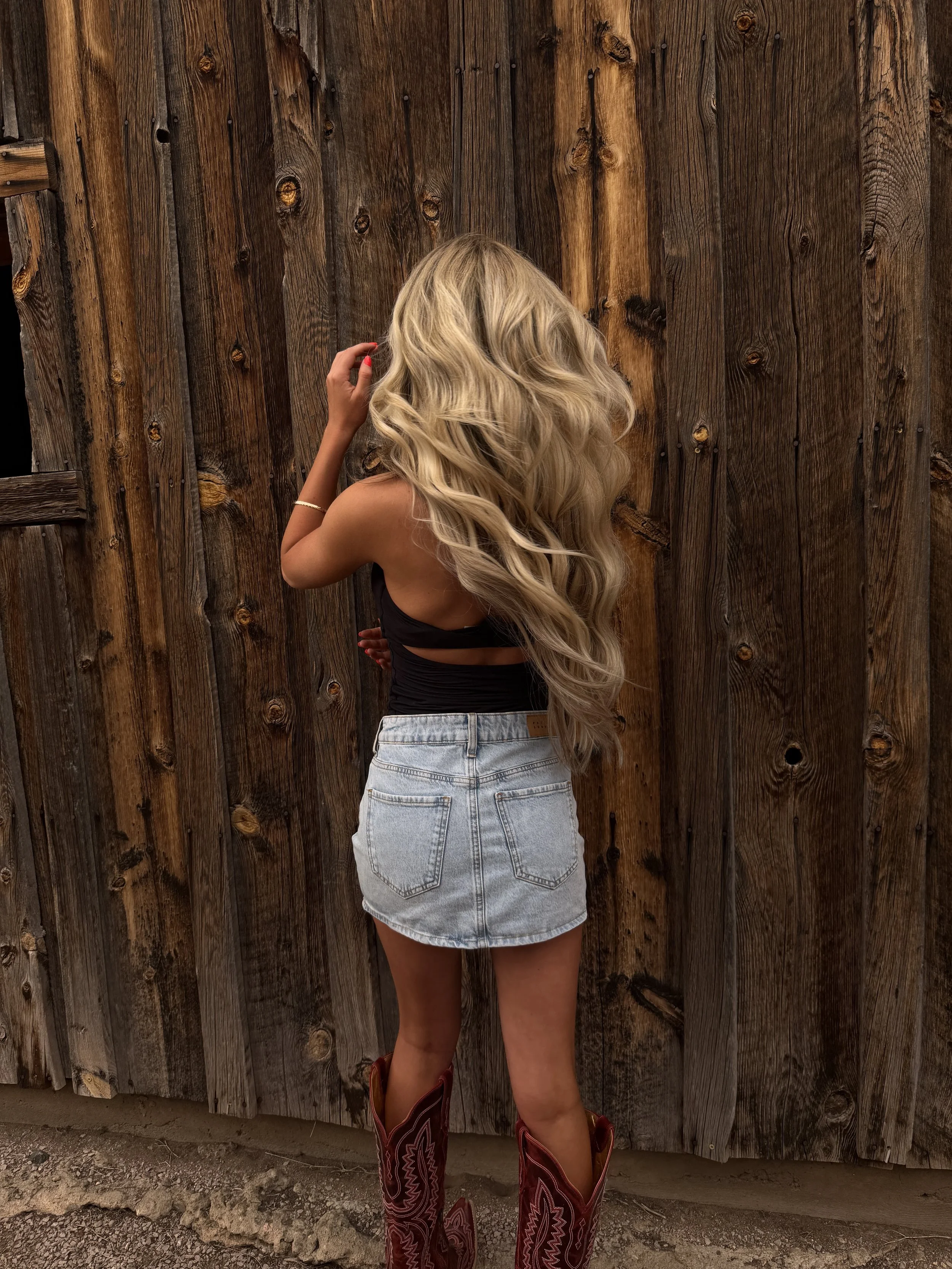 A woman with long, blonde, wavy hair is standing with her back to the camera in front of a wooden fence. She is wearing a black sleeveless top, high-waisted denim shorts, and cowboy boots.
