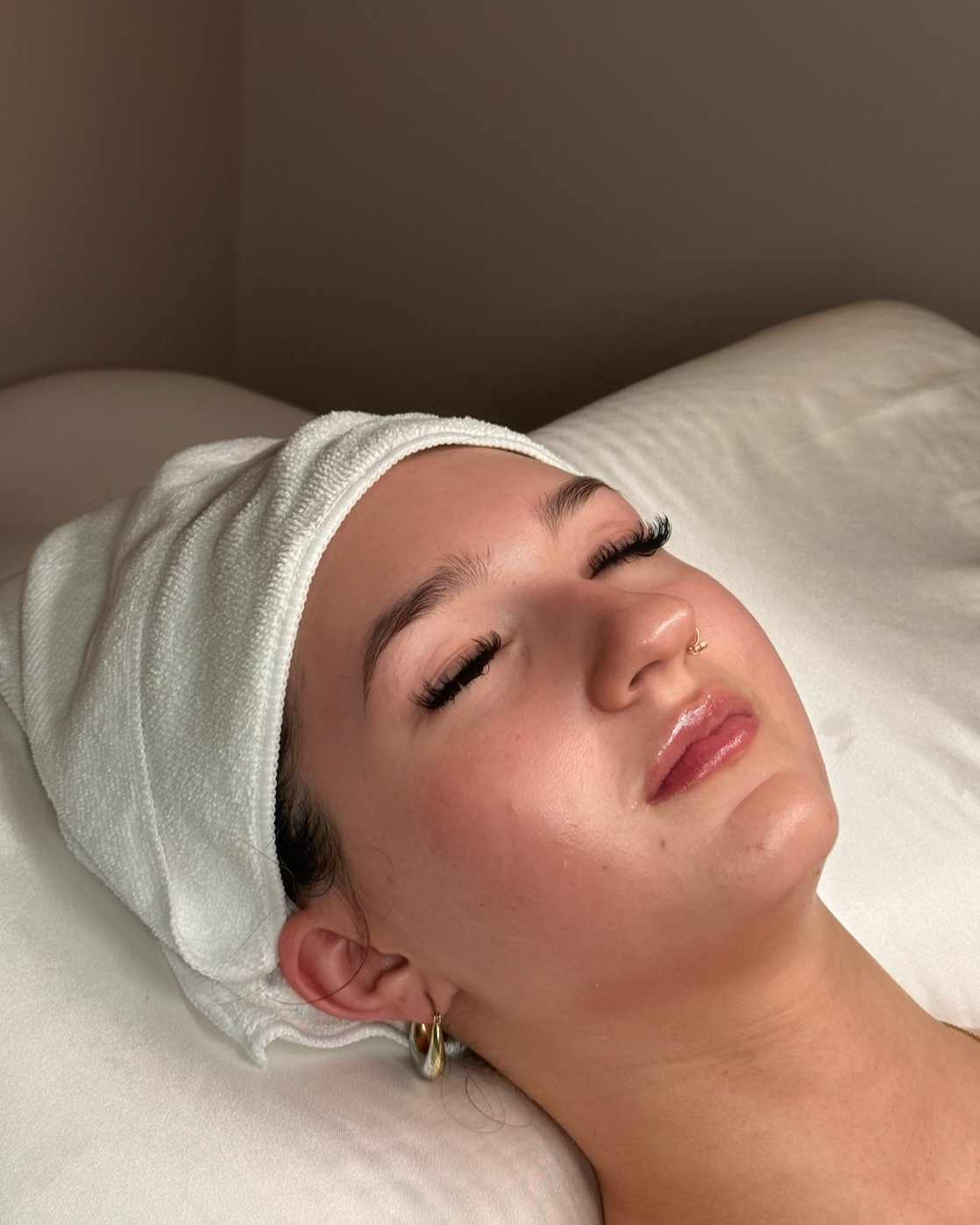 A woman lying on a bed with her eyes closed, wearing a white headband and hoop earrings, appearing relaxed.