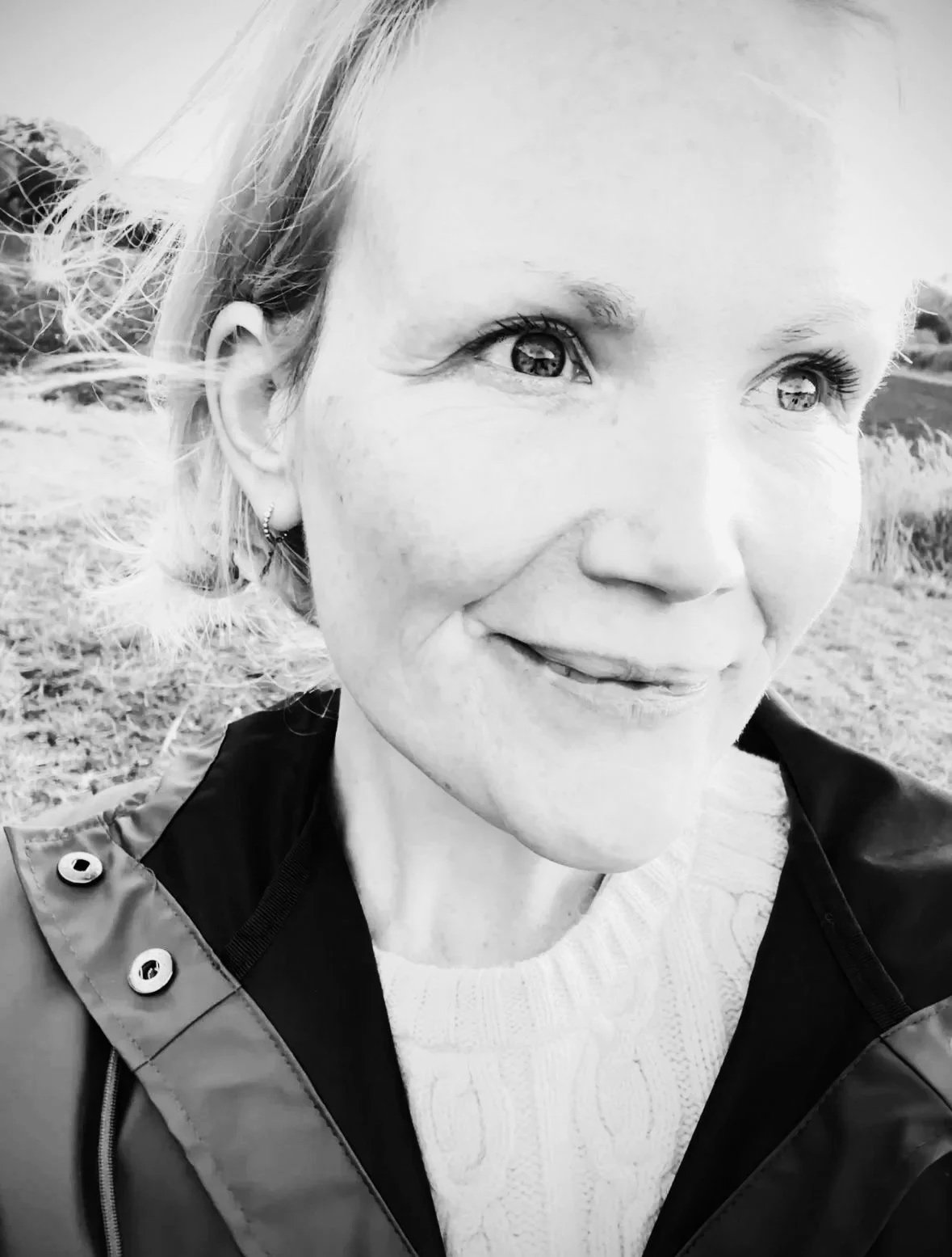 Close-up black and white photo of a smiling woman with light hair and an earring, outdoors in a natural setting.