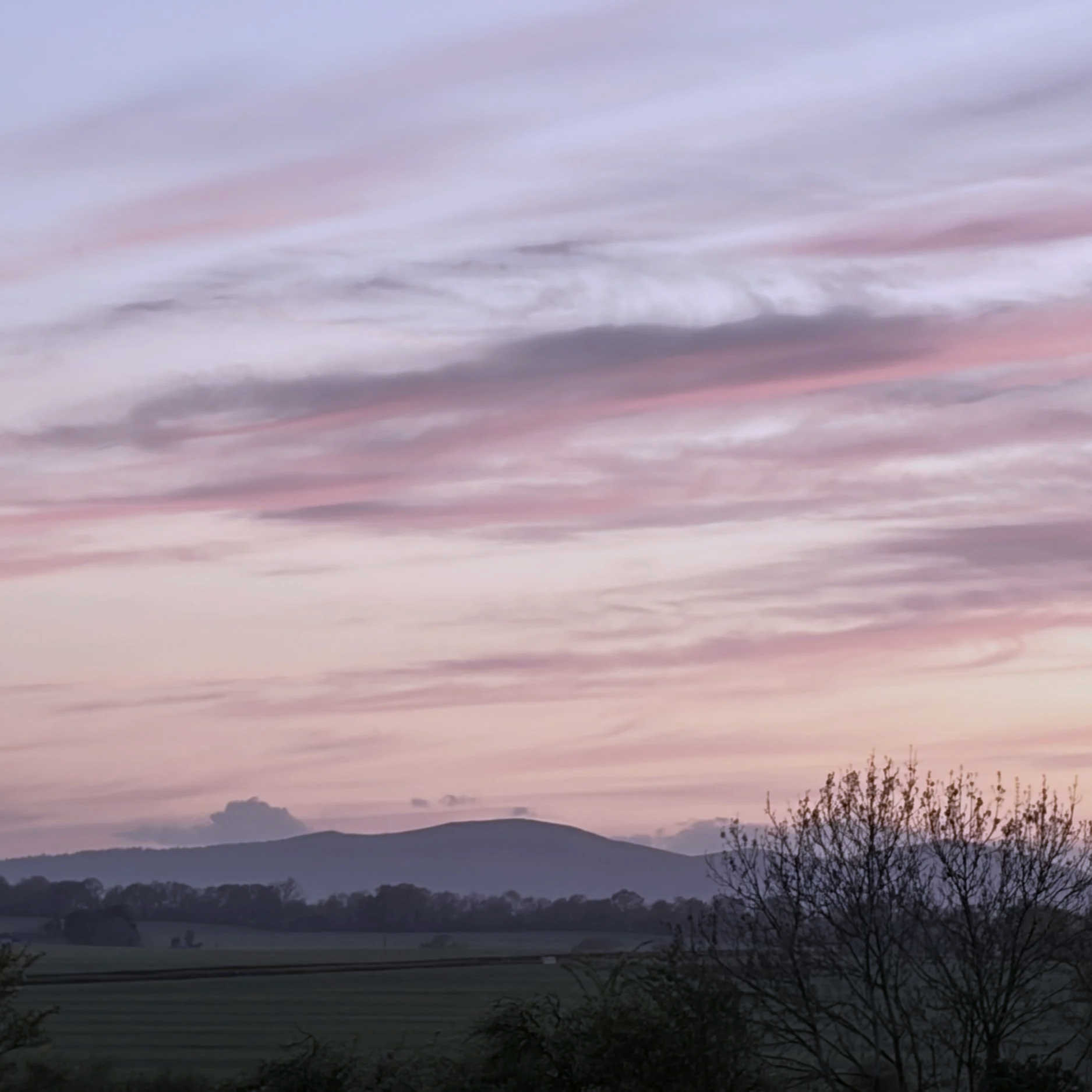 A landscape scene at dusk or dawn showing sky with streaks of pink, purple, and gray clouds above dark mountains and trees in the foreground.