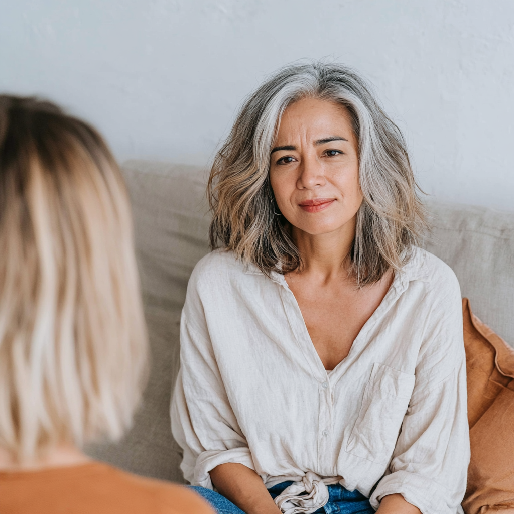 A middle-aged woman with gray, wavy hair sitting on a beige couch, looking at another woman with blonde hair, engaging in a serious conversation.