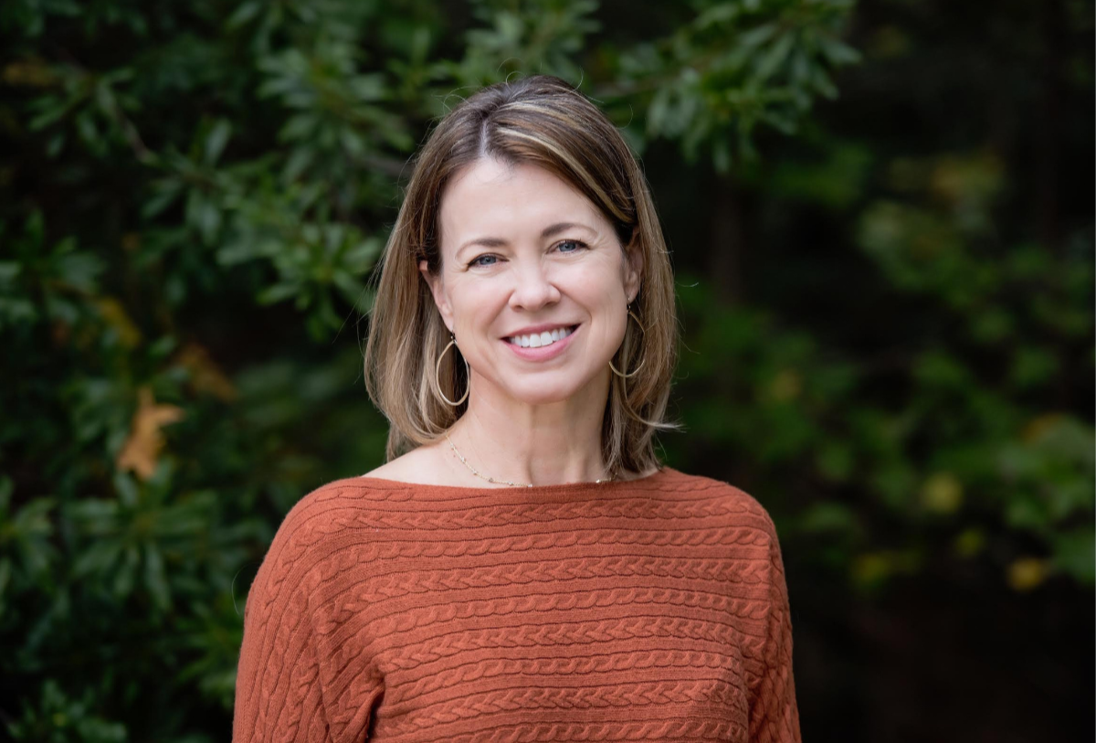 A woman with shoulder-length brown hair, wearing a rust-colored cable-knit sweater and hoop earrings, smiling outdoors with greenery in the background.