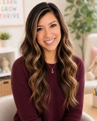 A smiling young woman with long, wavy brown hair, wearing a burgundy sweater, sitting in a cozy room with plants, a white chair, and a colorful framed poster in the background.
