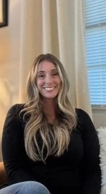Smiling woman with long blonde hair, wearing a black top, sitting indoors near a window with blinds.