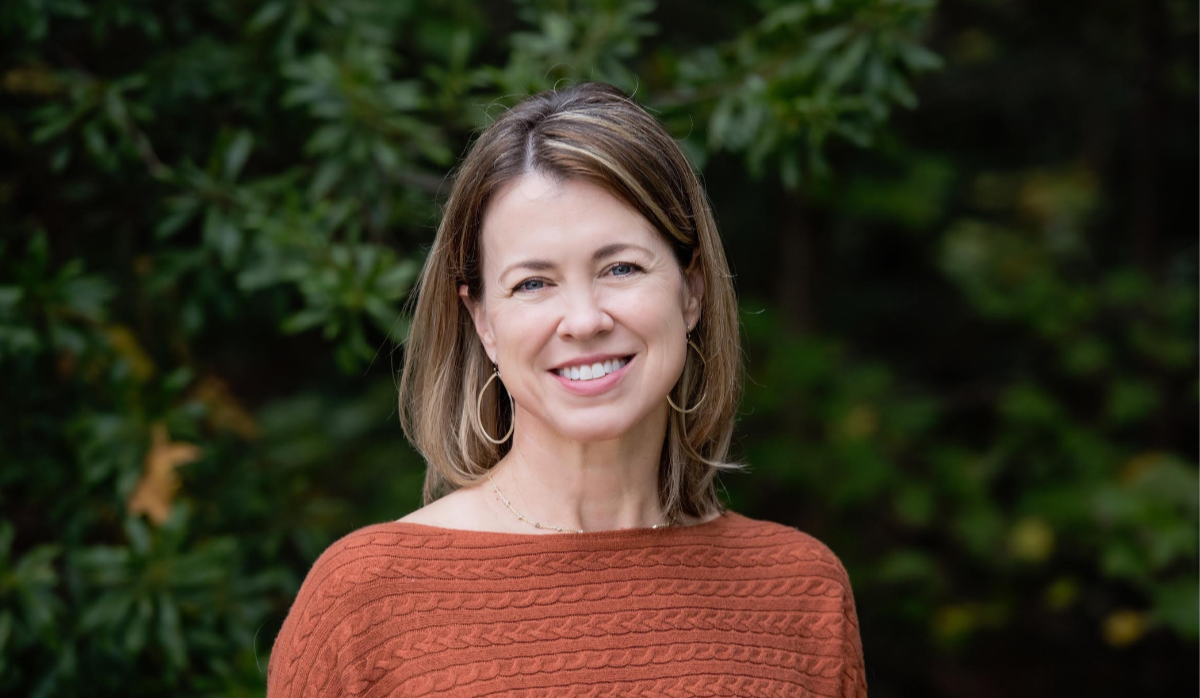 A woman with shoulder-length light brown hair, wearing hoop earrings and a rust-colored knitted sweater, standing outdoors with greenery in the background and smiling.