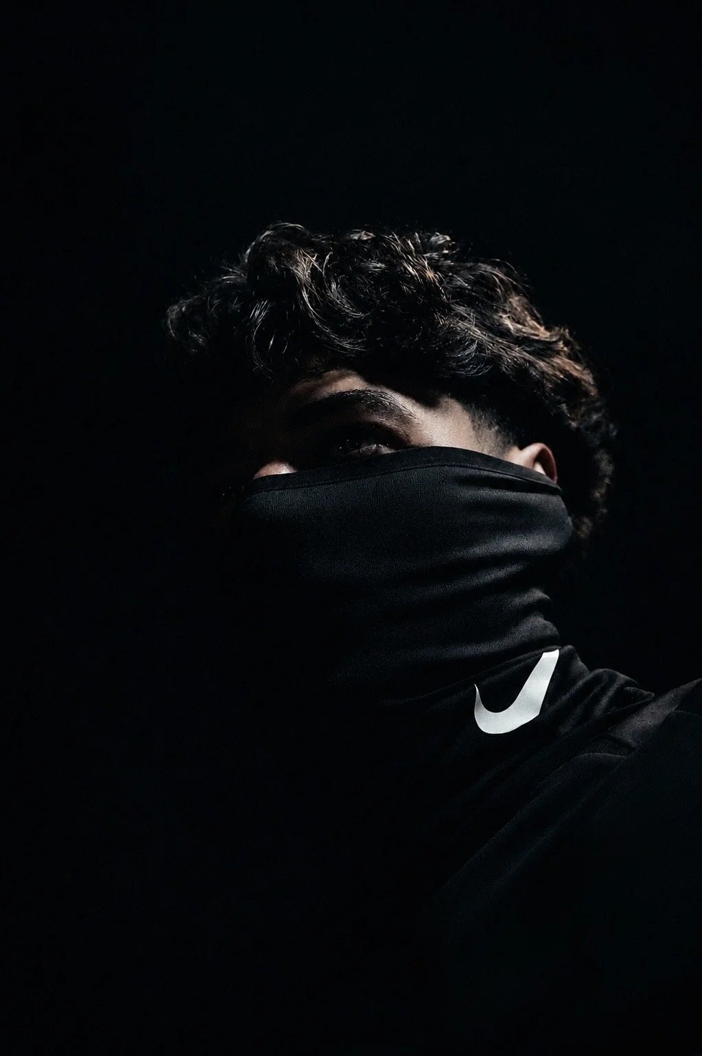 A person with curly hair wearing a black face mask and a black Nike shirt, partially illuminated in a dark setting.