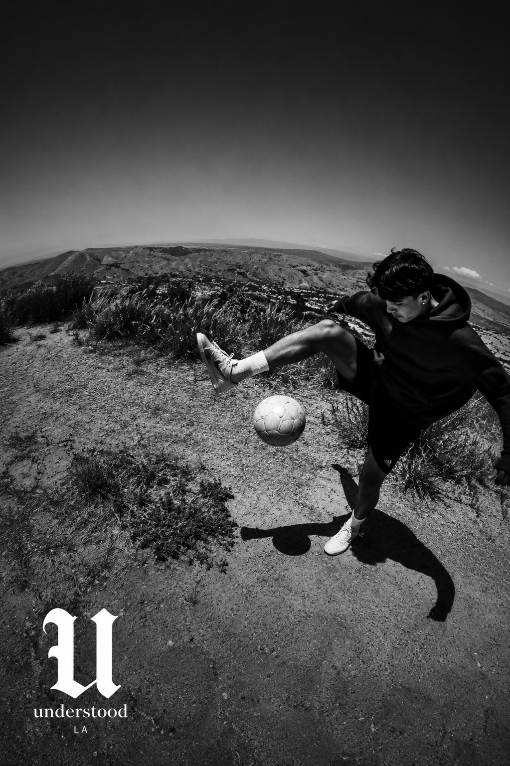 Black and white photograph of a young man in athletic gear kicking a soccer ball on a dirt trail with hilly terrain in the background and a clear sky.
