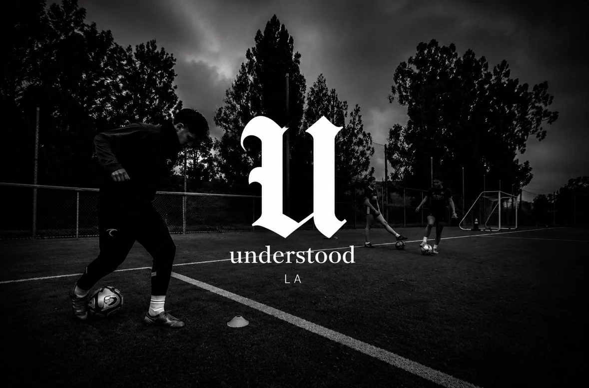 Black and white photo of young soccer players practicing on an outdoor field during cloudy weather, with trees in the background and a large newspaper-style logo in the center reading 'understood LA'.