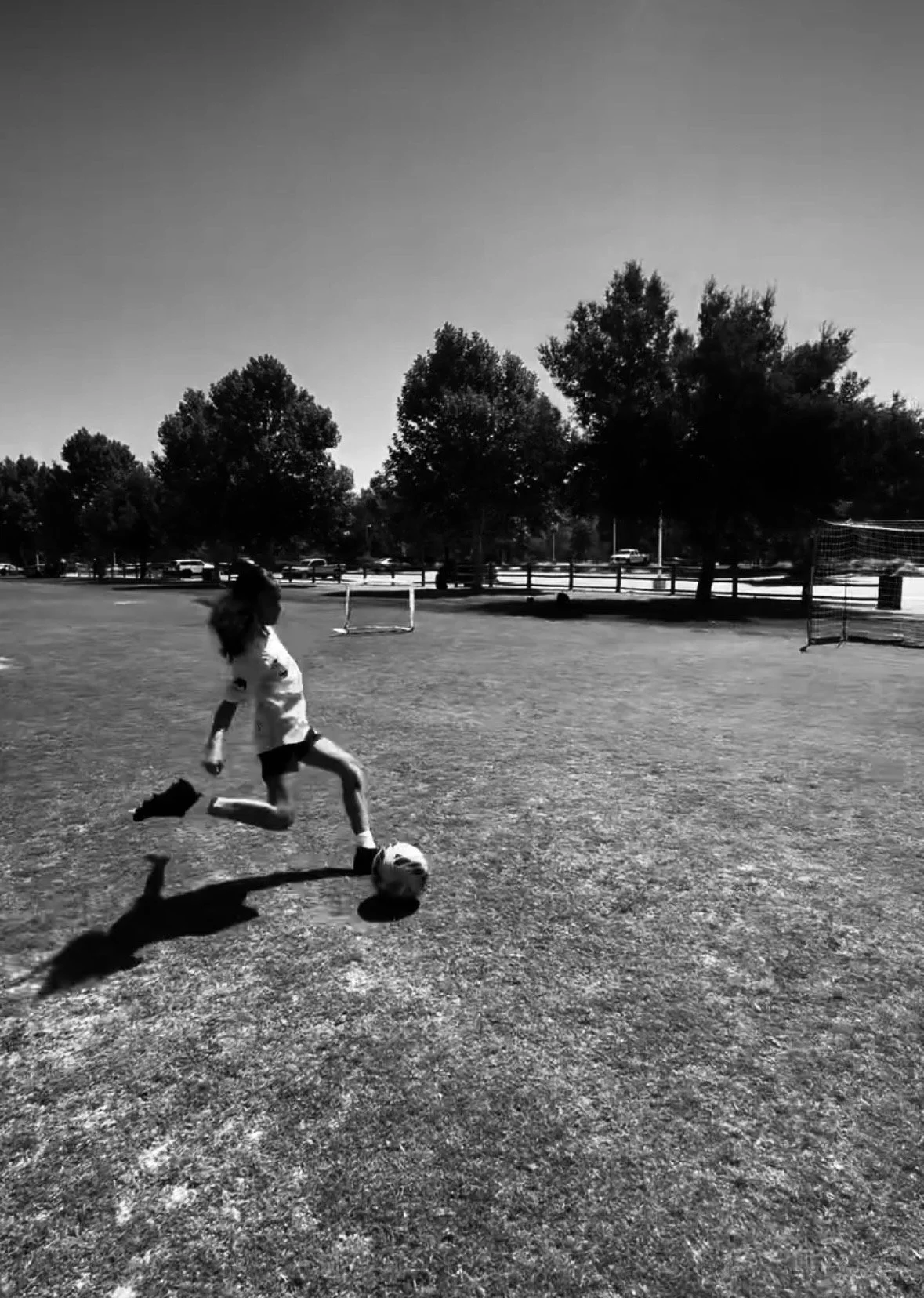 A girl playing soccer on a field with trees and a parking lot in the background, in black and white.