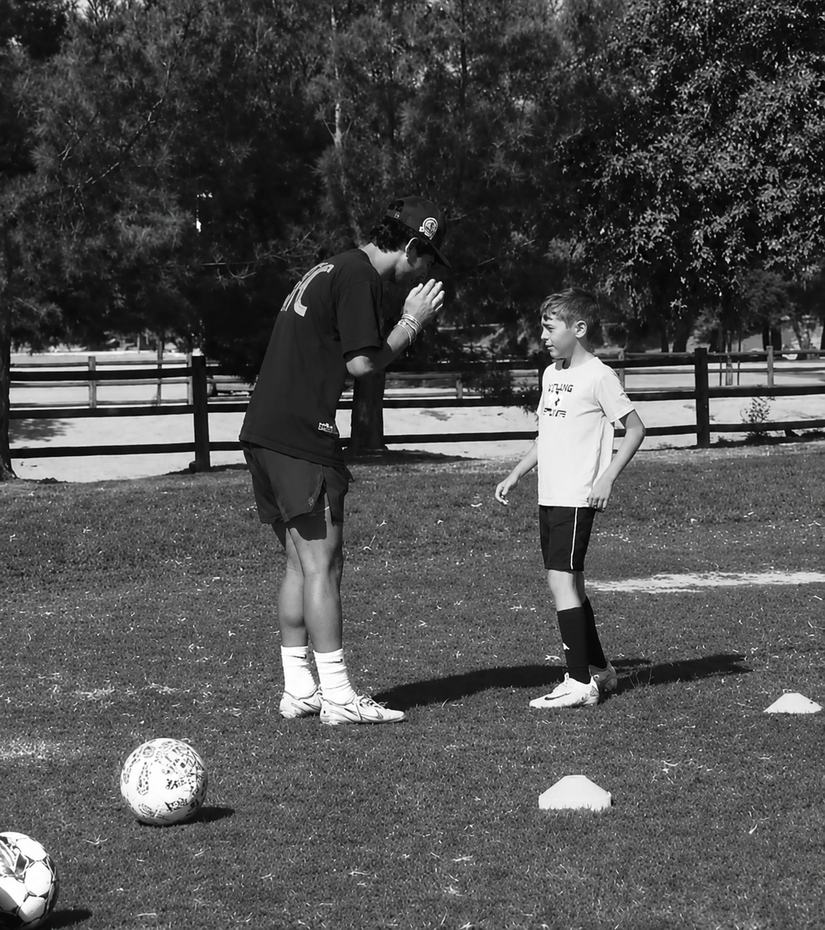 A coach and a young soccer player interact during practice on a grassy field, with soccer balls nearby and trees and a fence in the background.