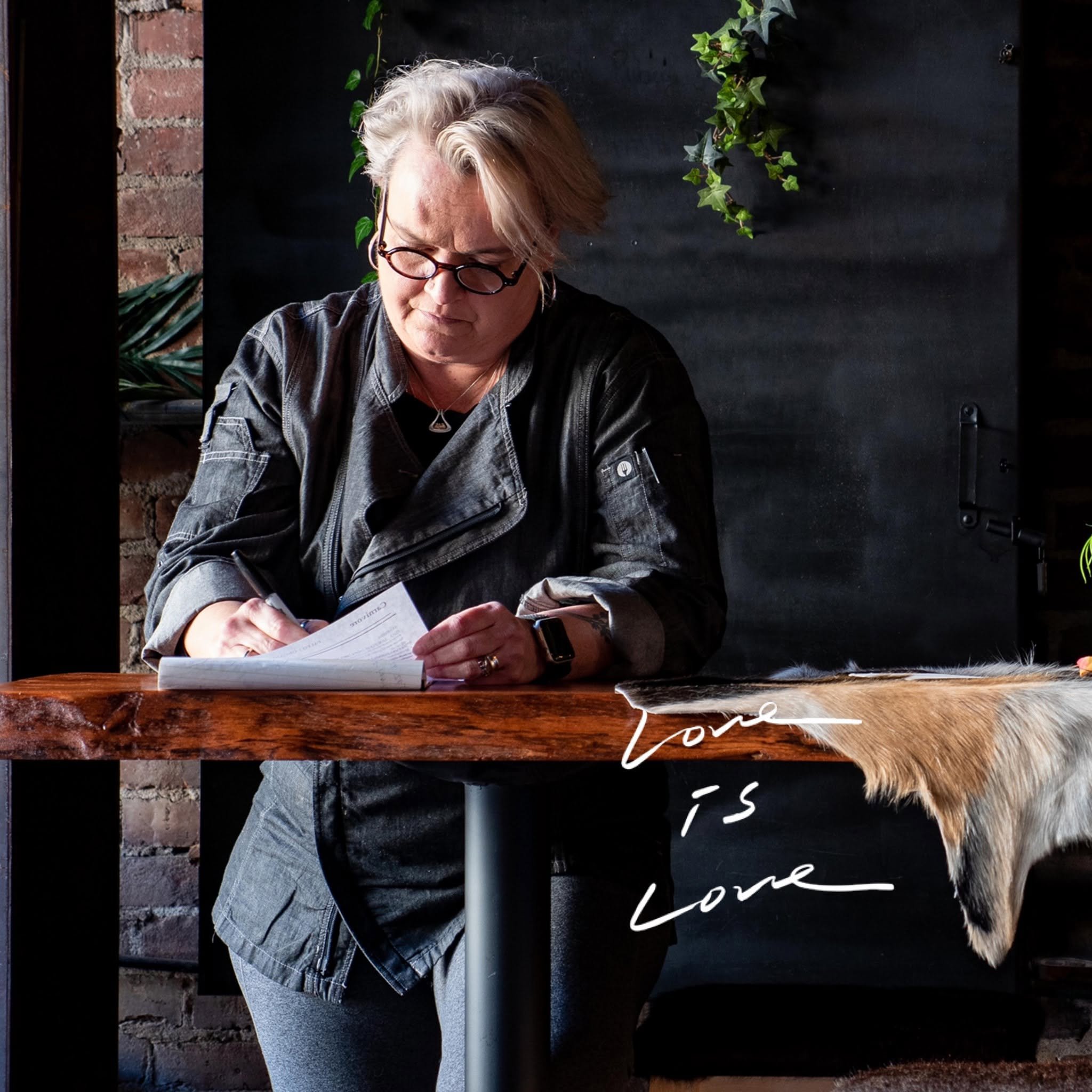A woman with gray hair and glasses sitting at a wooden table, reading a document, with green plants hanging on a black wall behind her.