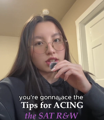 A young woman with long brown hair wearing glasses and a dark jacket, holding a snack near her mouth, in a home setting with a white door and beige walls.