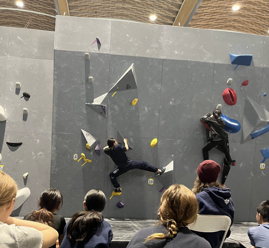 Two people rock climbing on an indoor climbing wall with multiple holds. Several spectators watching from below.