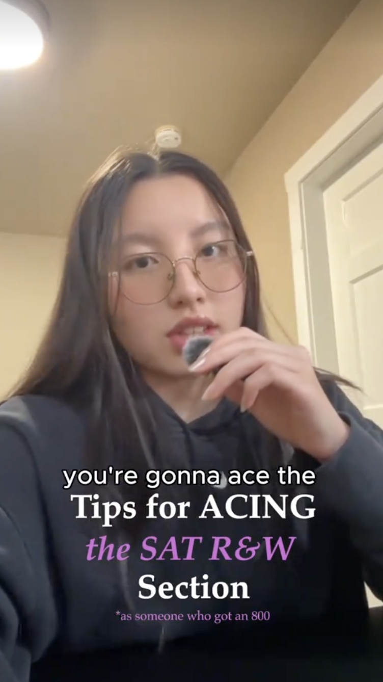 A young woman with long dark hair and glasses holding a pen near her mouth, standing indoors with a beige ceiling, a light fixture, and a white door visible behind her.