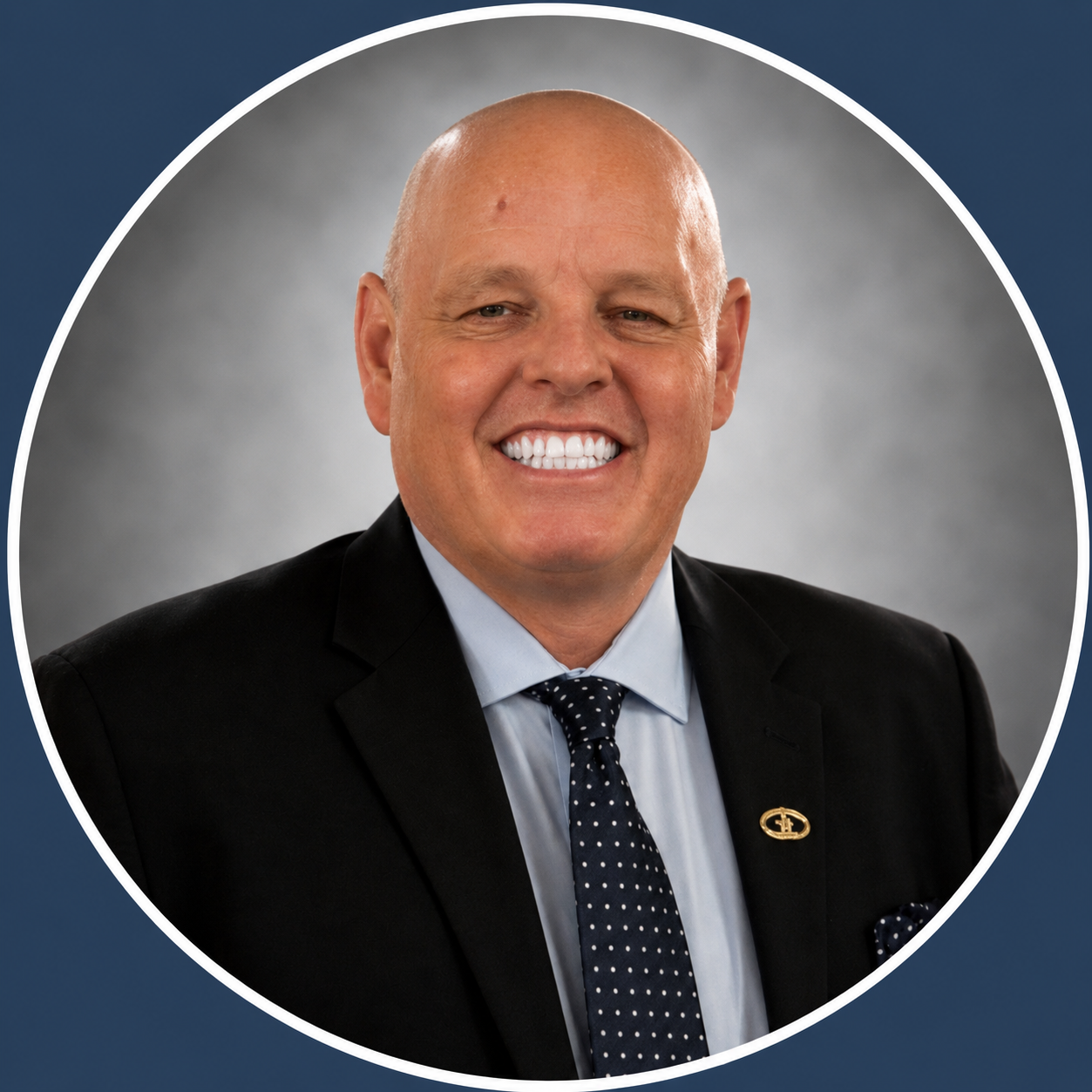 A professionally dressed man with a broad smile, wearing a black suit, light blue shirt, navy blue polka dot tie, and a lapel pin, posing against a gray background.