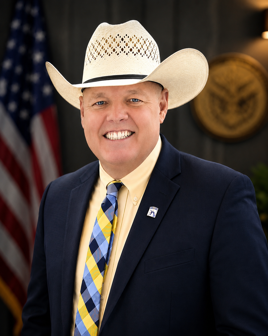 A man wearing a large white cowboy hat, a yellow dress shirt, a dark navy suit, and a blue, yellow, and white plaid tie. He is smiling and standing in front of a dark background with an American flag and a circular emblem.