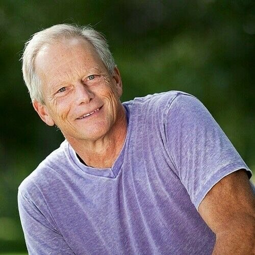 A smiling elderly man with gray hair wearing a purple T-shirt outdoors with green foliage in the background.