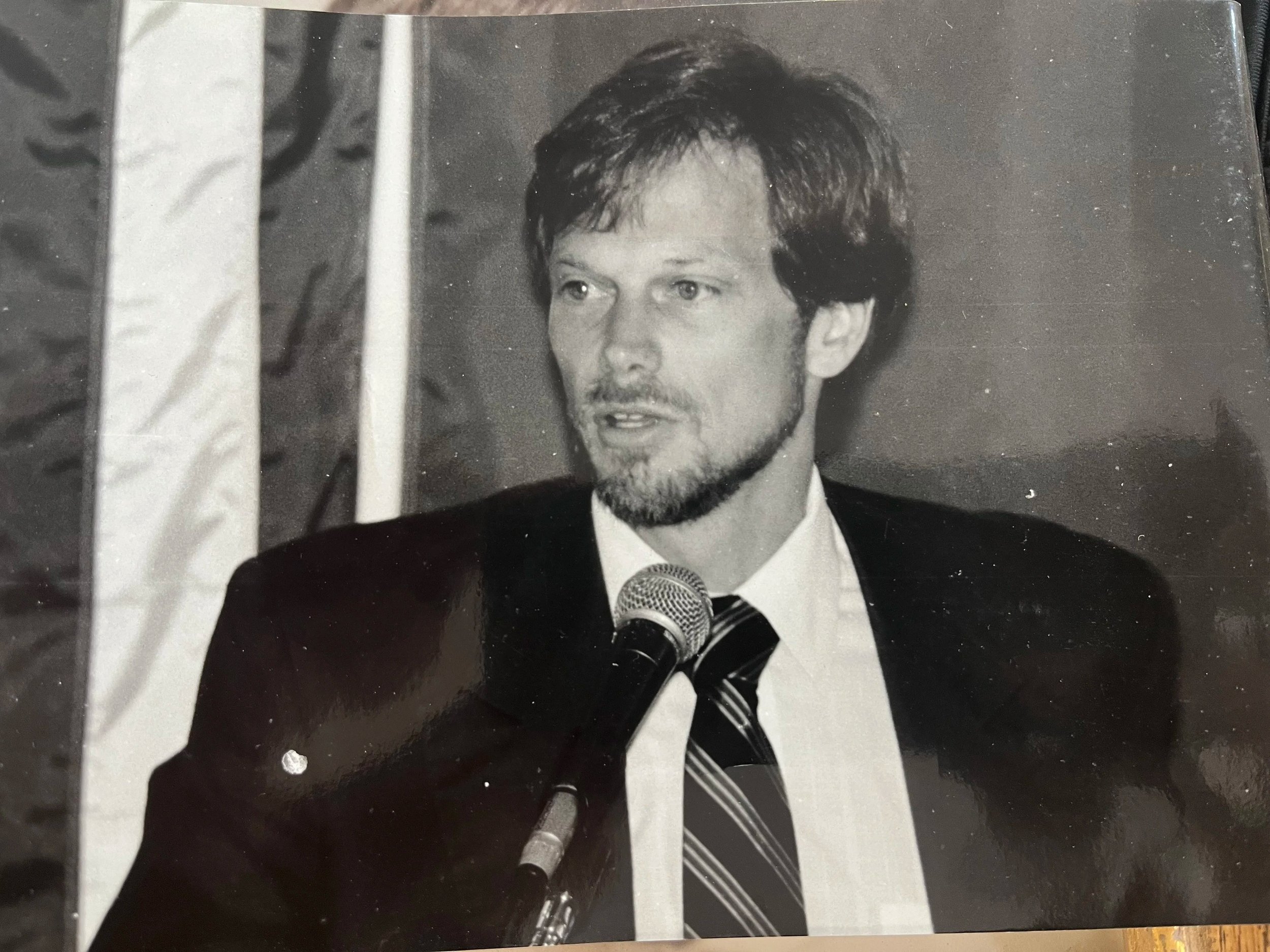 A black and white photograph of a man with dark hair and a beard, dressed in a suit and tie, speaking into a microphone