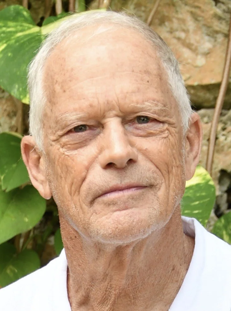 Close-up of an elderly man with white hair, light skin, and blue eyes, standing outdoors with green plants in the background.