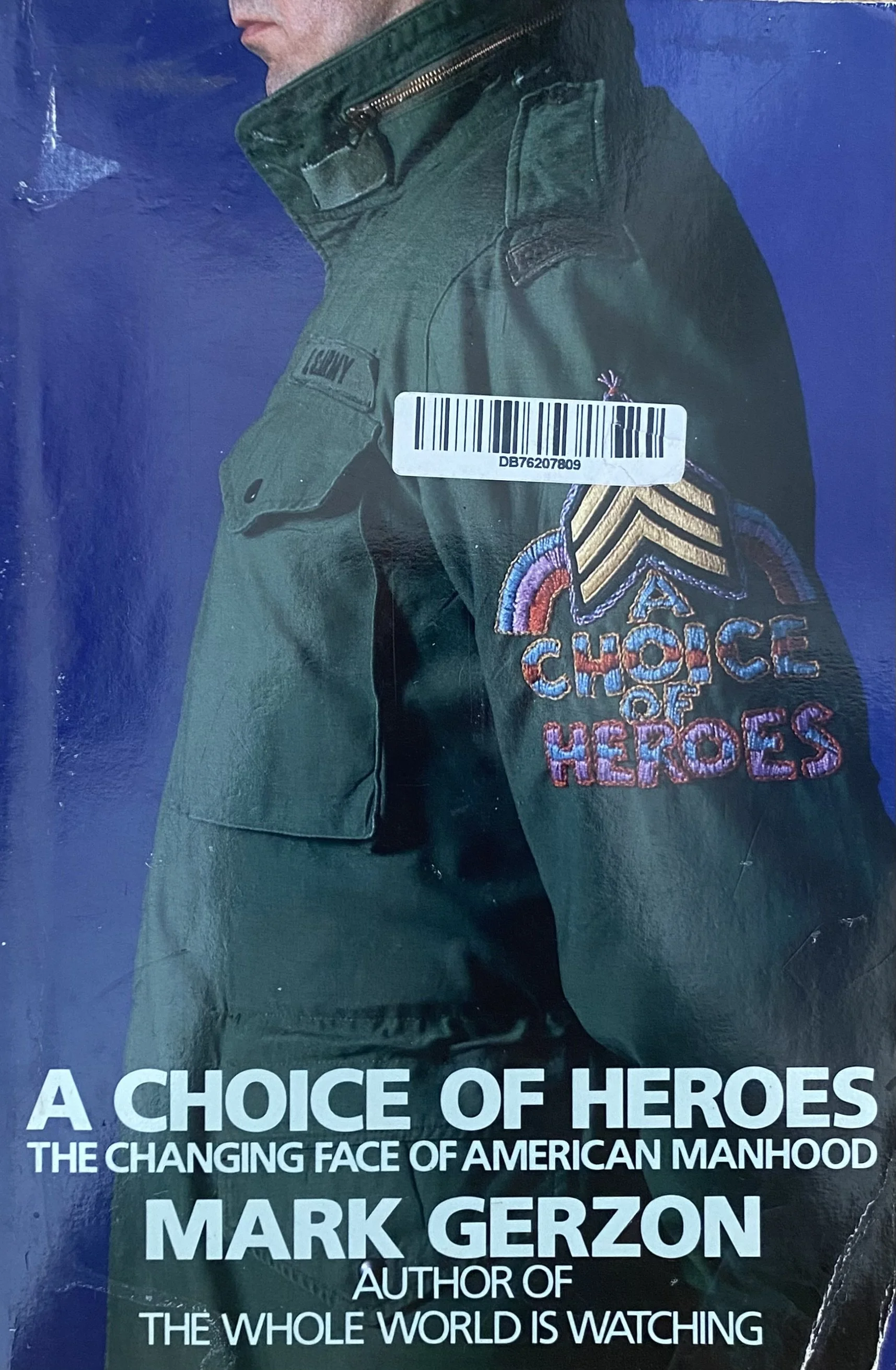 Close-up of a man in a green military jacket with a patch that reads 'LOYALTY' on the arm. The jacket has a dramatic embroidered patch with rainbow and chevron designs that says "CHOOSE HEROES." The background is a plain blue wall, and the bottom of the image features text about the book titled 'A CHOICE OF HEROES' by Mark Gerzon.