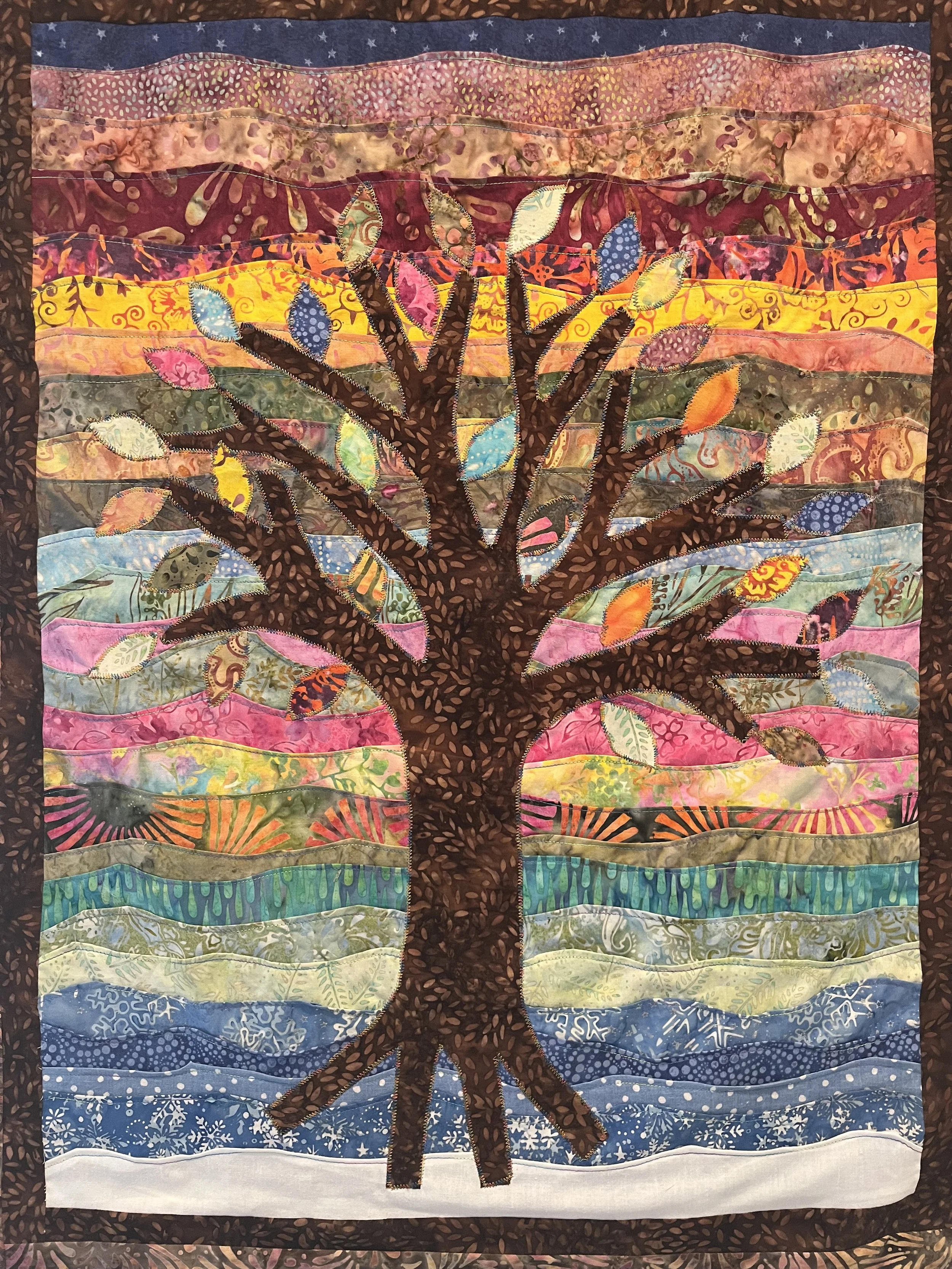A quilt depicting a colorful tree with a dark brown trunk and branches, set against a background of layered, multicolored fabric resembling a landscape with rolling hills and a sky in shades of pink, purple, yellow, blue, and gray. The quilt is framed with a dark brown border with leaf-like patterns.
