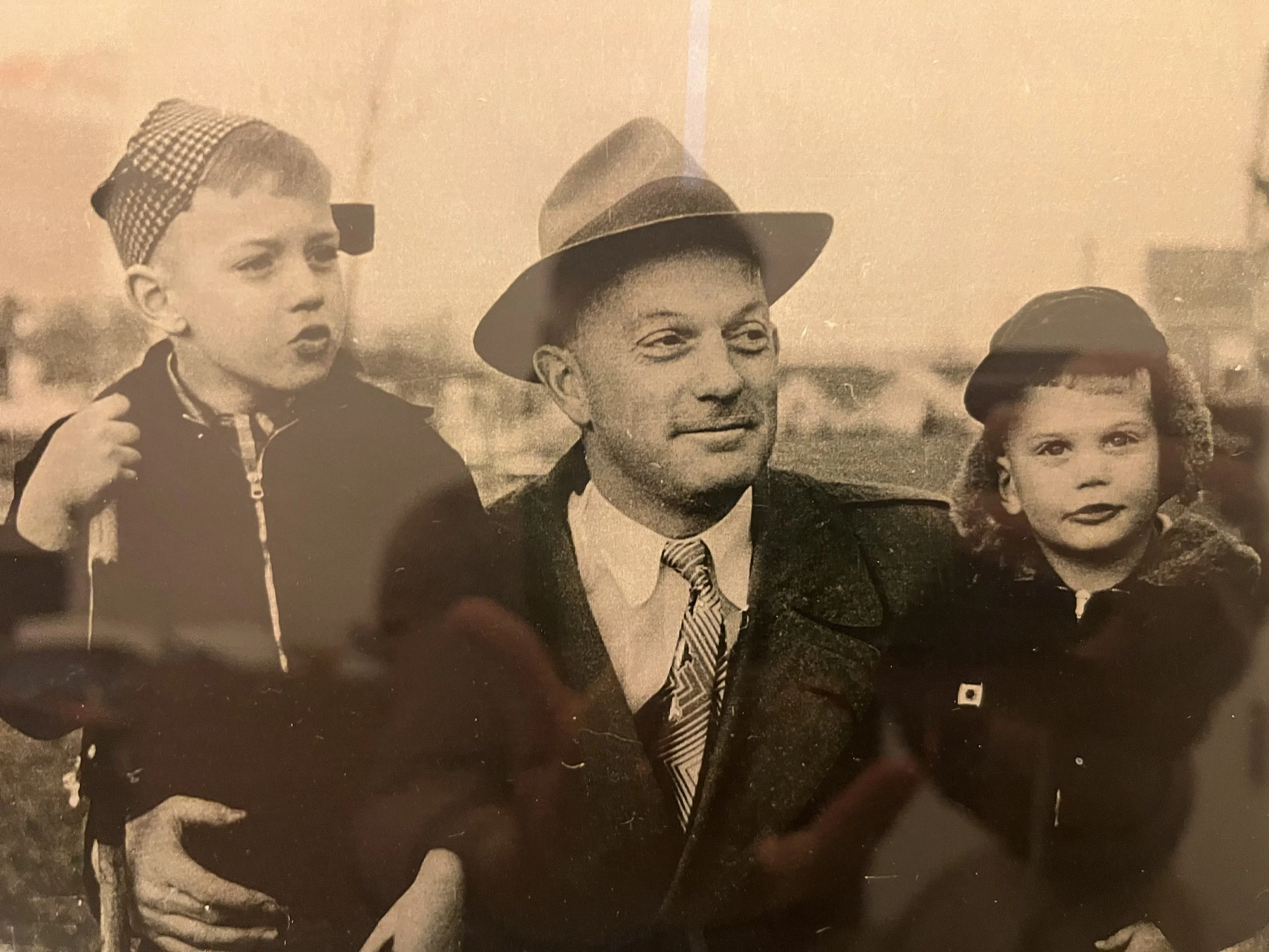 A black-and-white photo of a man wearing a fedora and a woman and a young girl, all dressed in vintage clothing, standing outdoors with a blurred background.