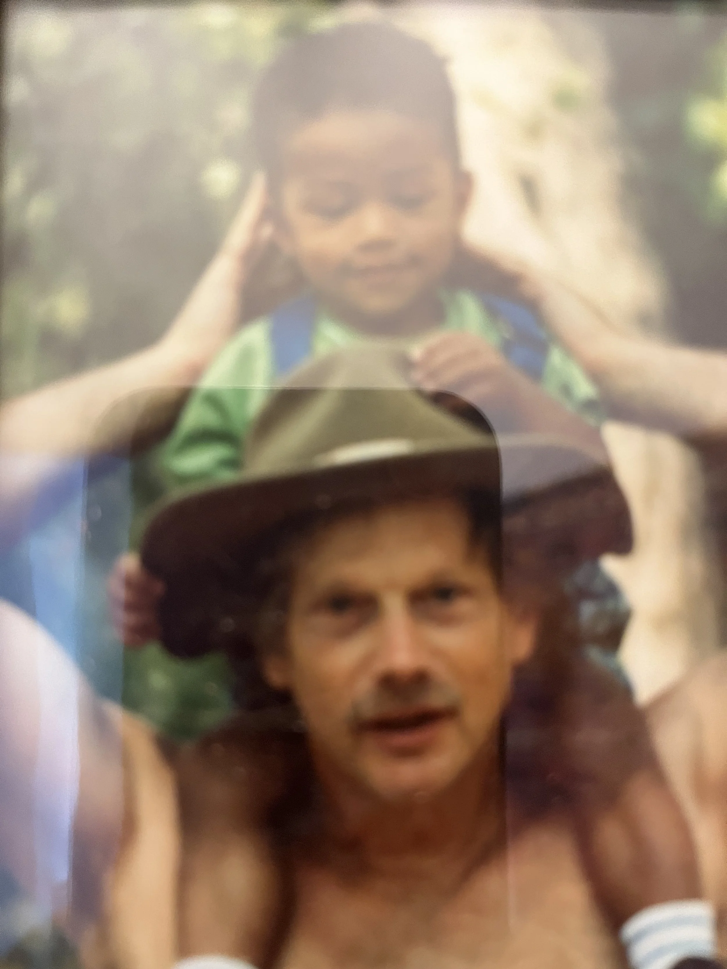 A man with dark hair and a mustache wearing a cap, and a young boy with short dark hair, headphones, and colorful clothing, outdoors in a natural setting.