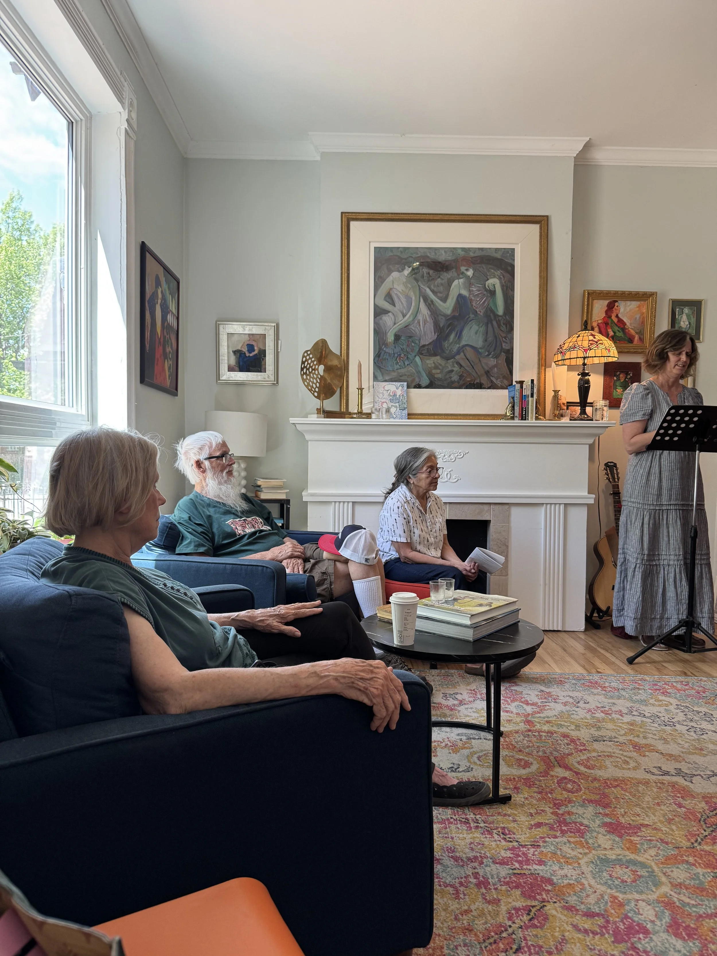 A group of people attending a gathering in a living room, with artwork on the walls, a large window letting in natural light, and a woman standing at a music stand reading. The seated attendees include an elderly man with glasses and a white beard, a woman with glasses and a woman with gray hair, all sitting on sofas or chairs. There are books, a coffee cup, and a guitar visible in the room.