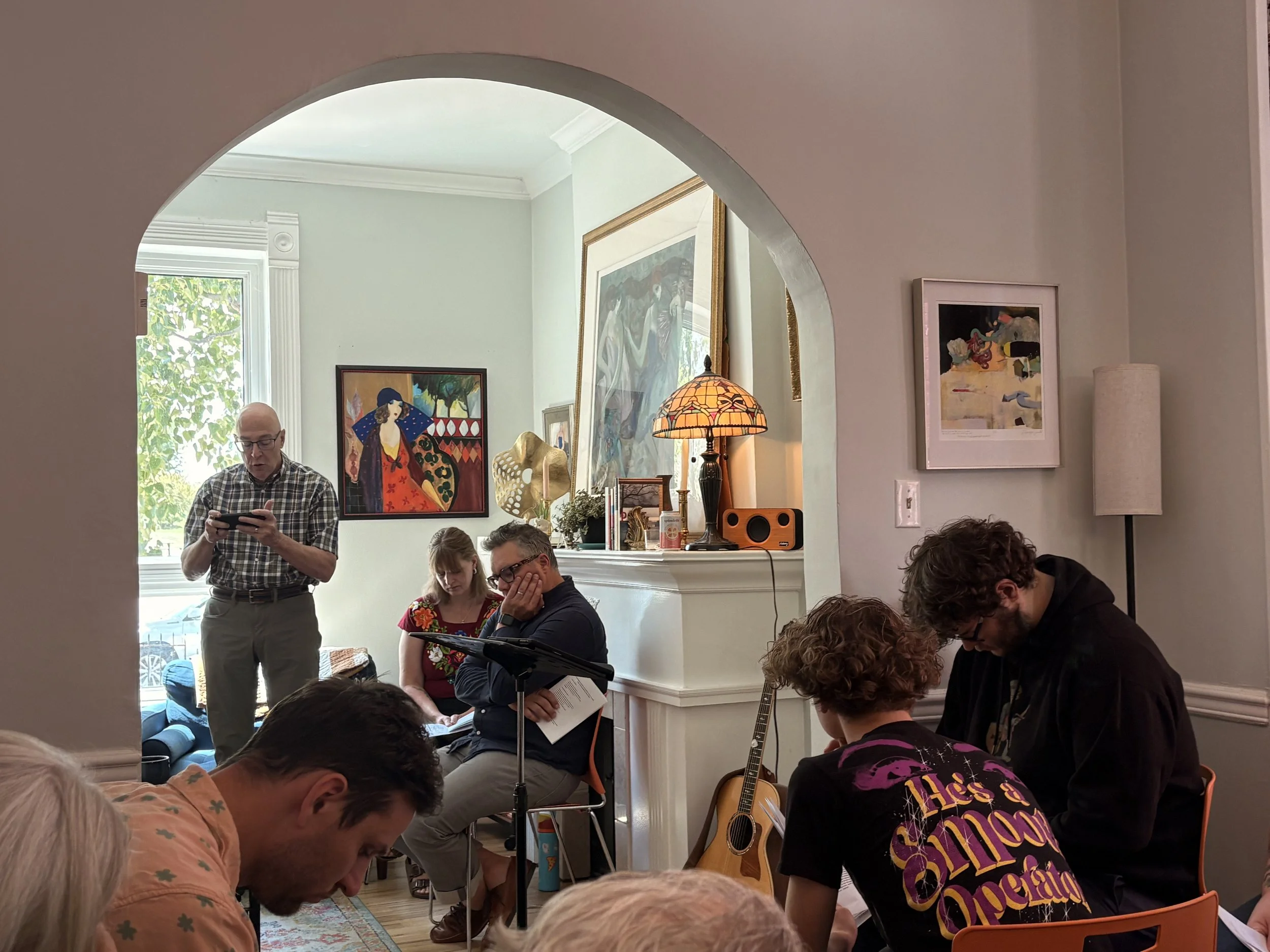 A group of people gathered indoors, some sitting and some standing, with artwork on the walls and a window with natural light. One person is standing, reading Scripture, while others listen.