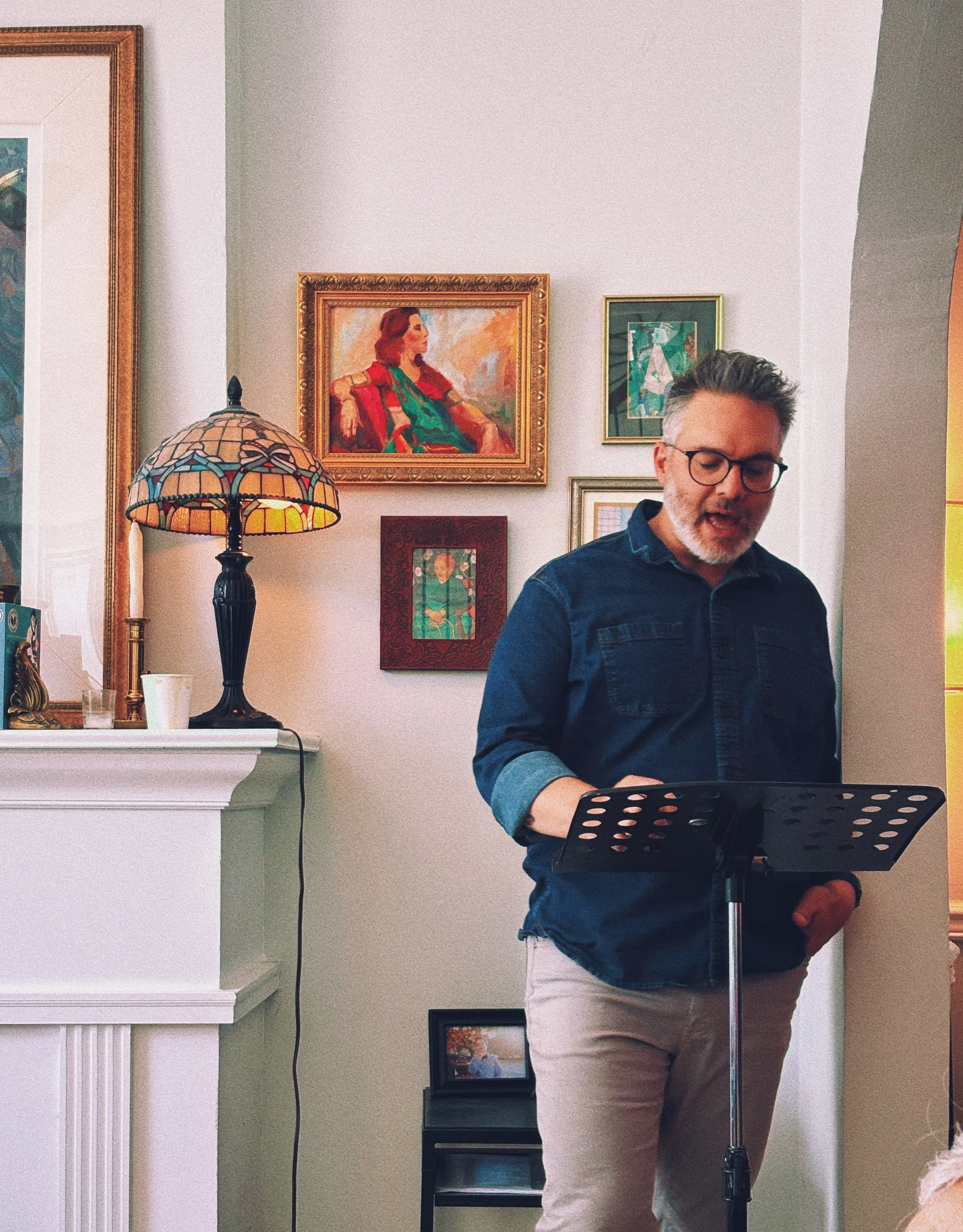 A man with glasses and gray hair, wearing a dark blue shirt and light-colored pants, is standing in front of a music stand, possibly singing or speaking, in a room with white walls decorated with framed artwork.