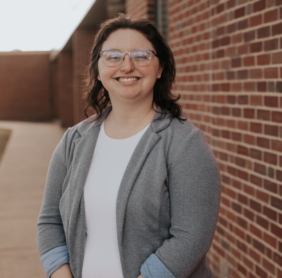 A woman with curly dark hair, glasses, and a big smile standing outdoors next to a brick wall.