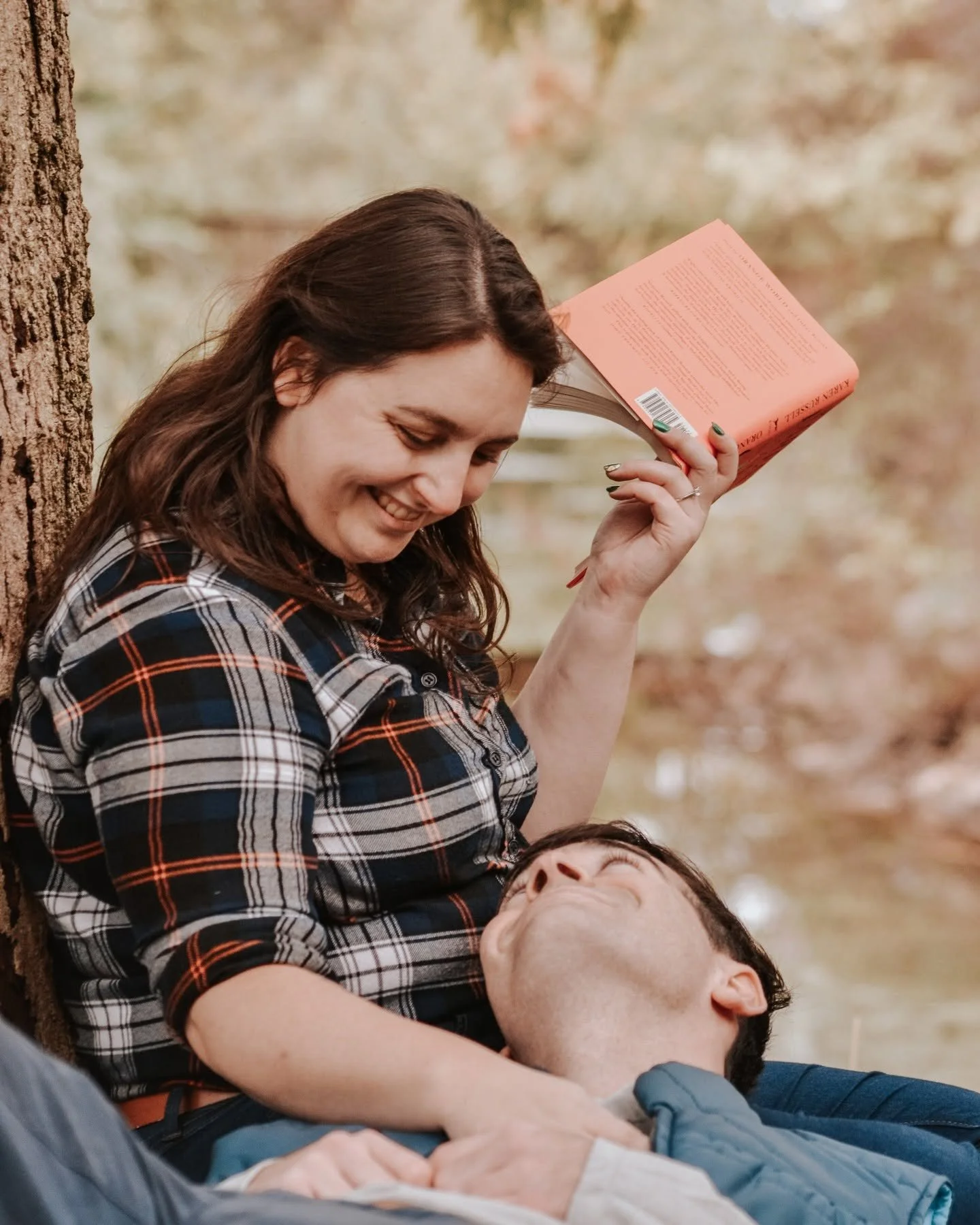 A woman with brown hair is smiling and leaning against a tree, holding a pink book near her head while looking down at a man lying on the ground, who is smiling with his eyes closed.