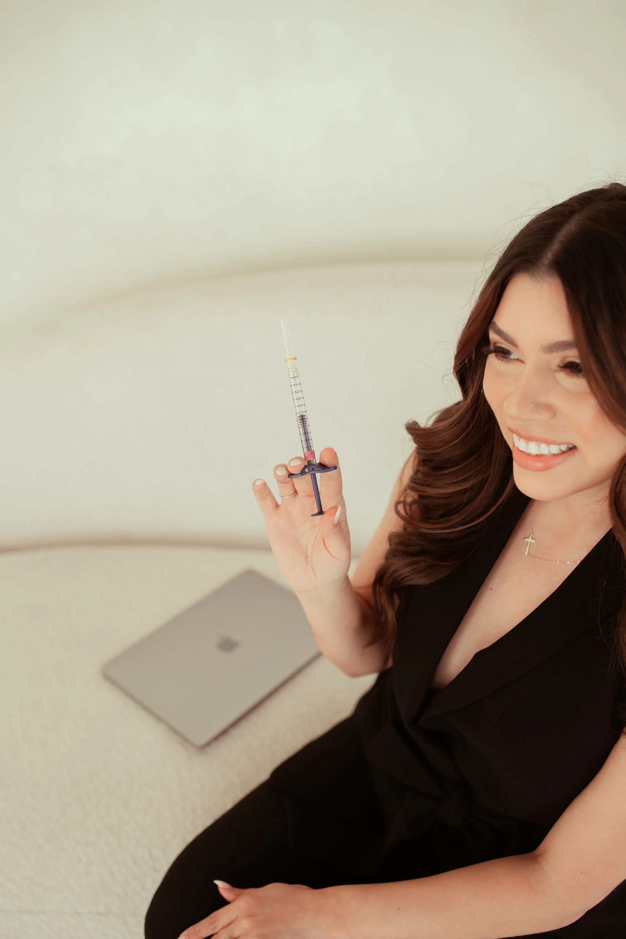 A woman smiling and holding a syringe while sitting at a table, in a black sleeveless top and a cross necklace.