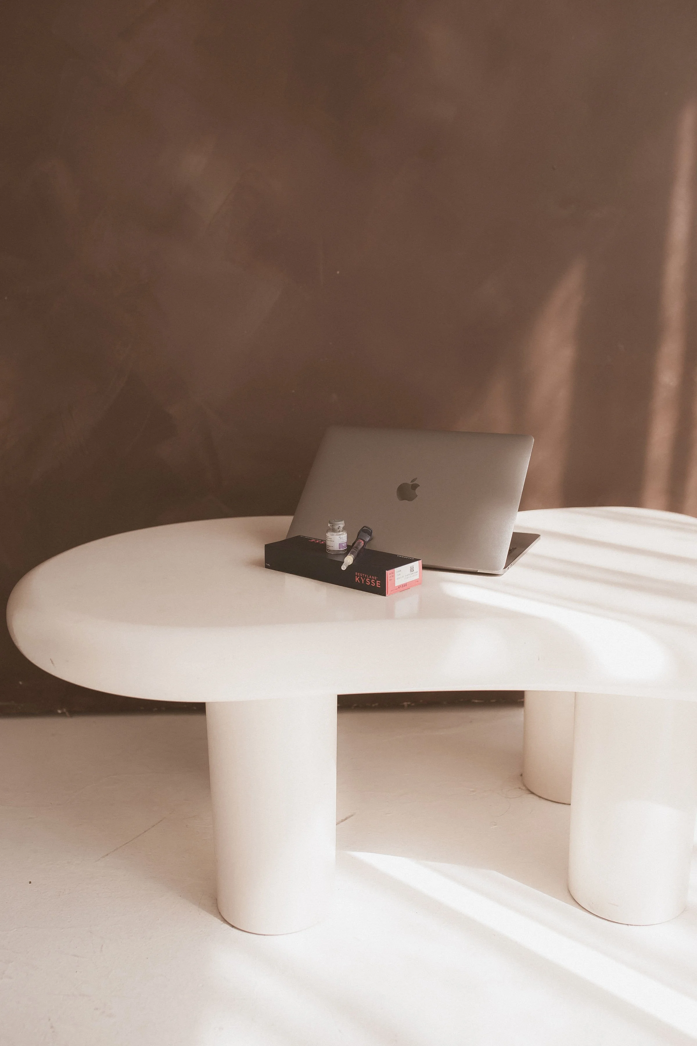 A round white table with cylindrical legs, a closed silver Apple laptop, a small bottle, and a black box with red and white labels on it are on the table. The background is a dark brown wall.