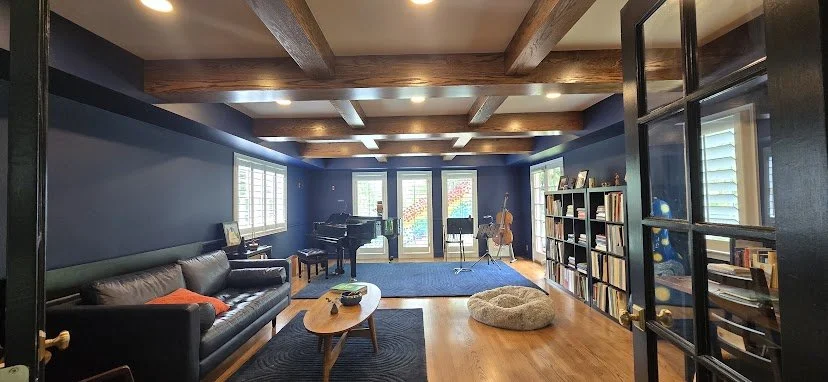 Spacious living room with a black sofa, coffee table, grand piano, bookshelves, and musical instruments, brightened by large windows and wooden ceiling beams.