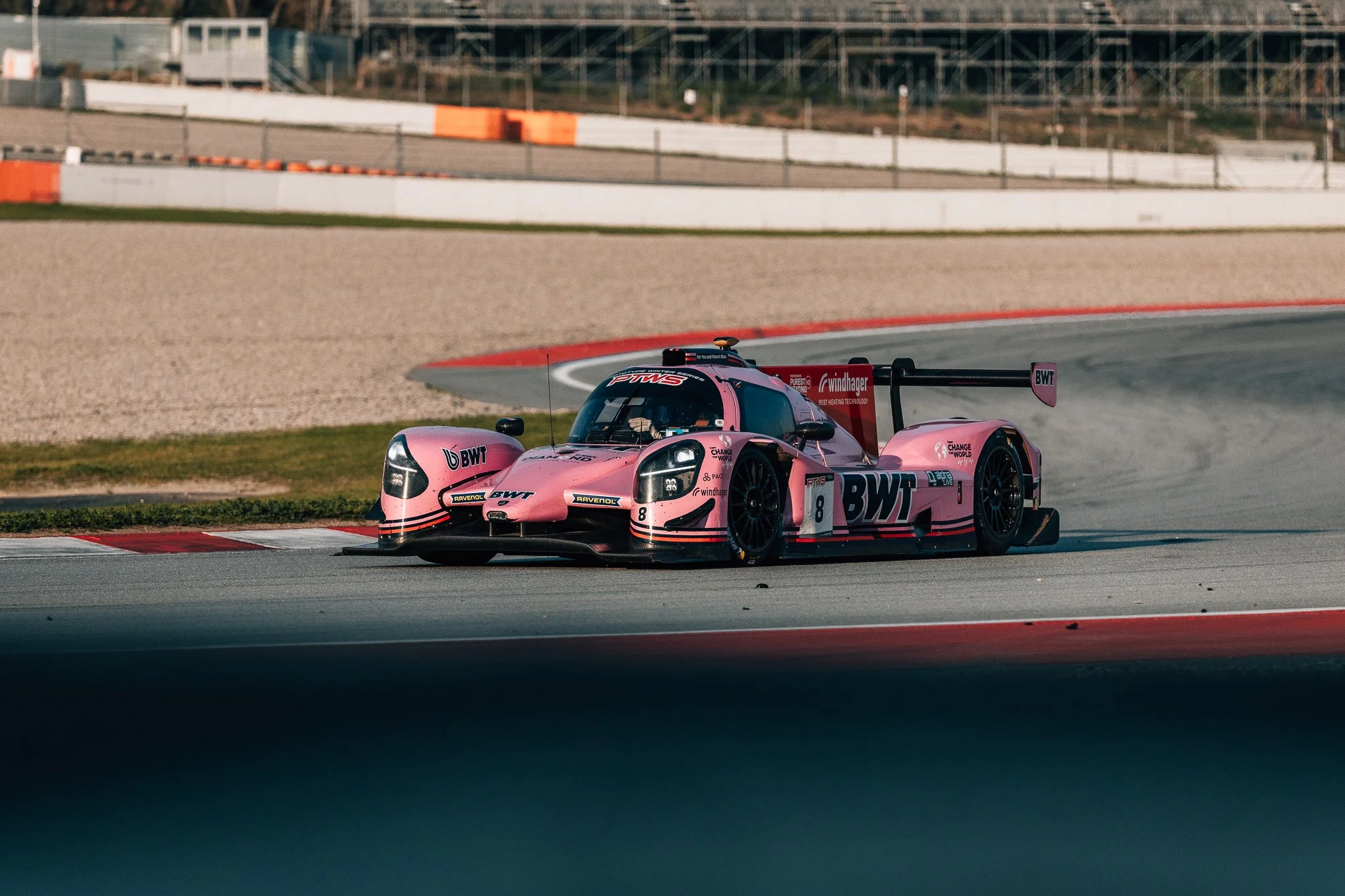A pink racing car on a racetrack during daytime, with barriers and structures in the background.