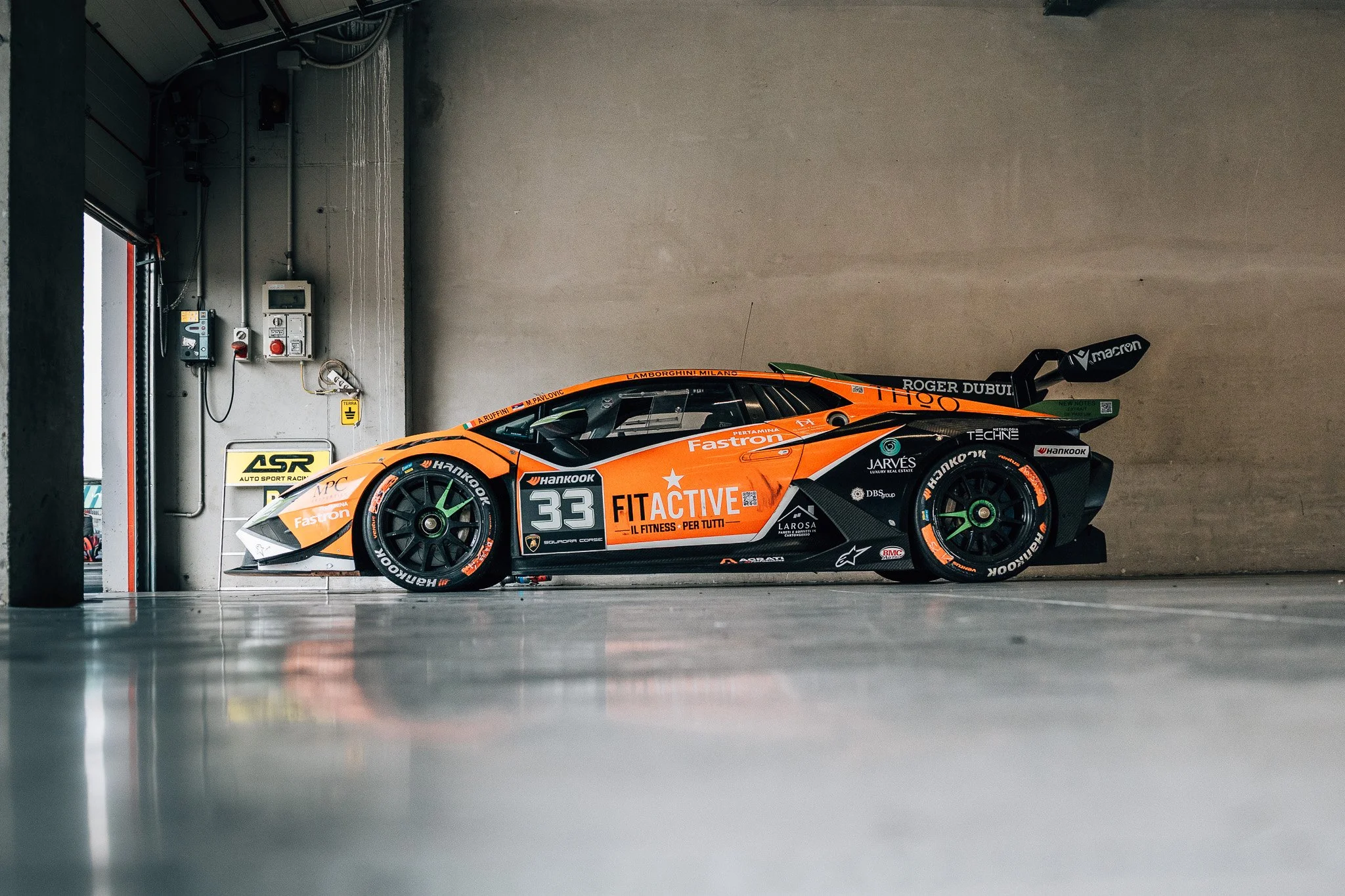 An orange race car with black accents and sponsorship logos, parked inside a garage. The car has the number 33 on the side and is positioned against a plain wall with various electrical equipment and signs.