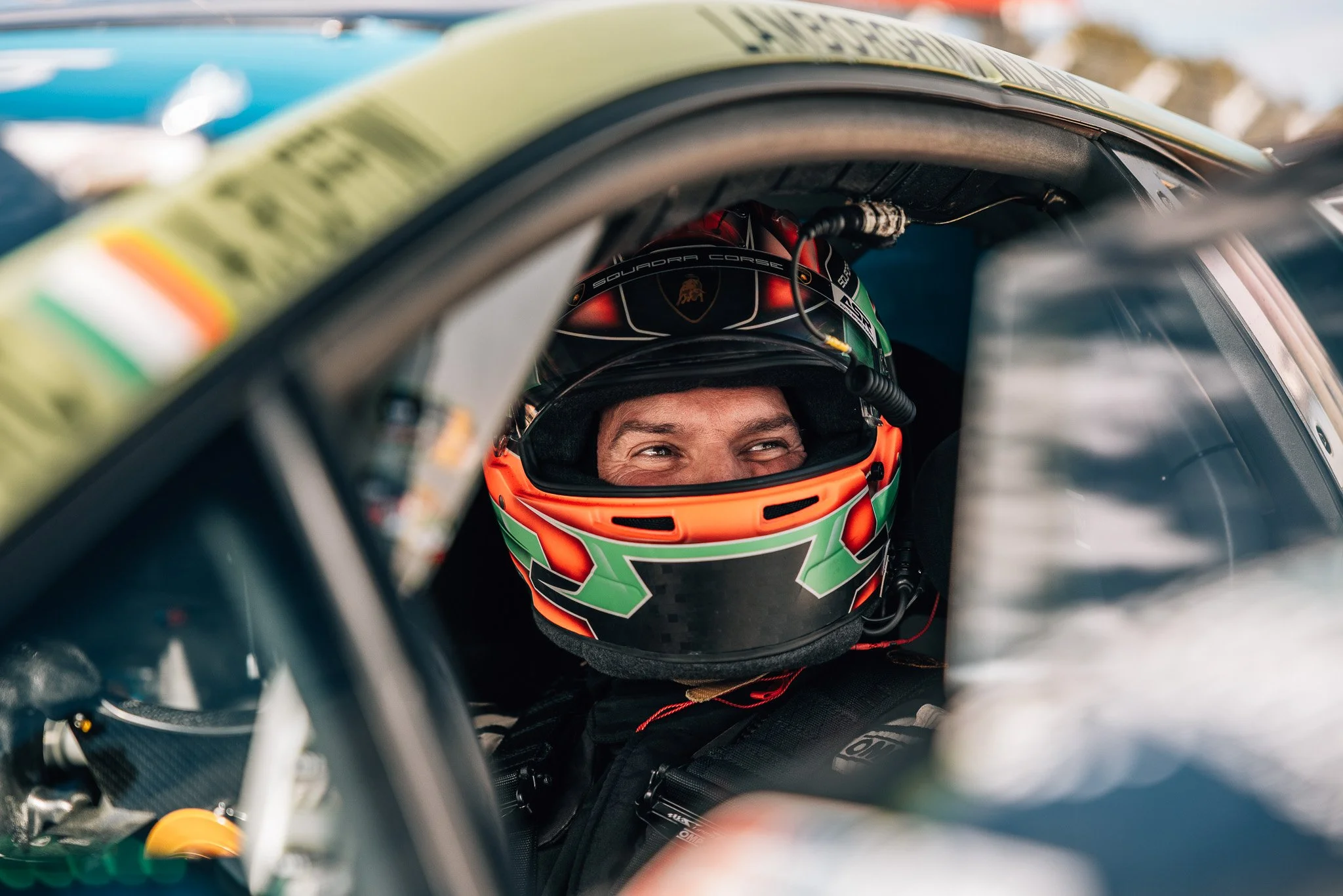 Close-up of a race car driver wearing a racing helmet, seated inside a racing car.