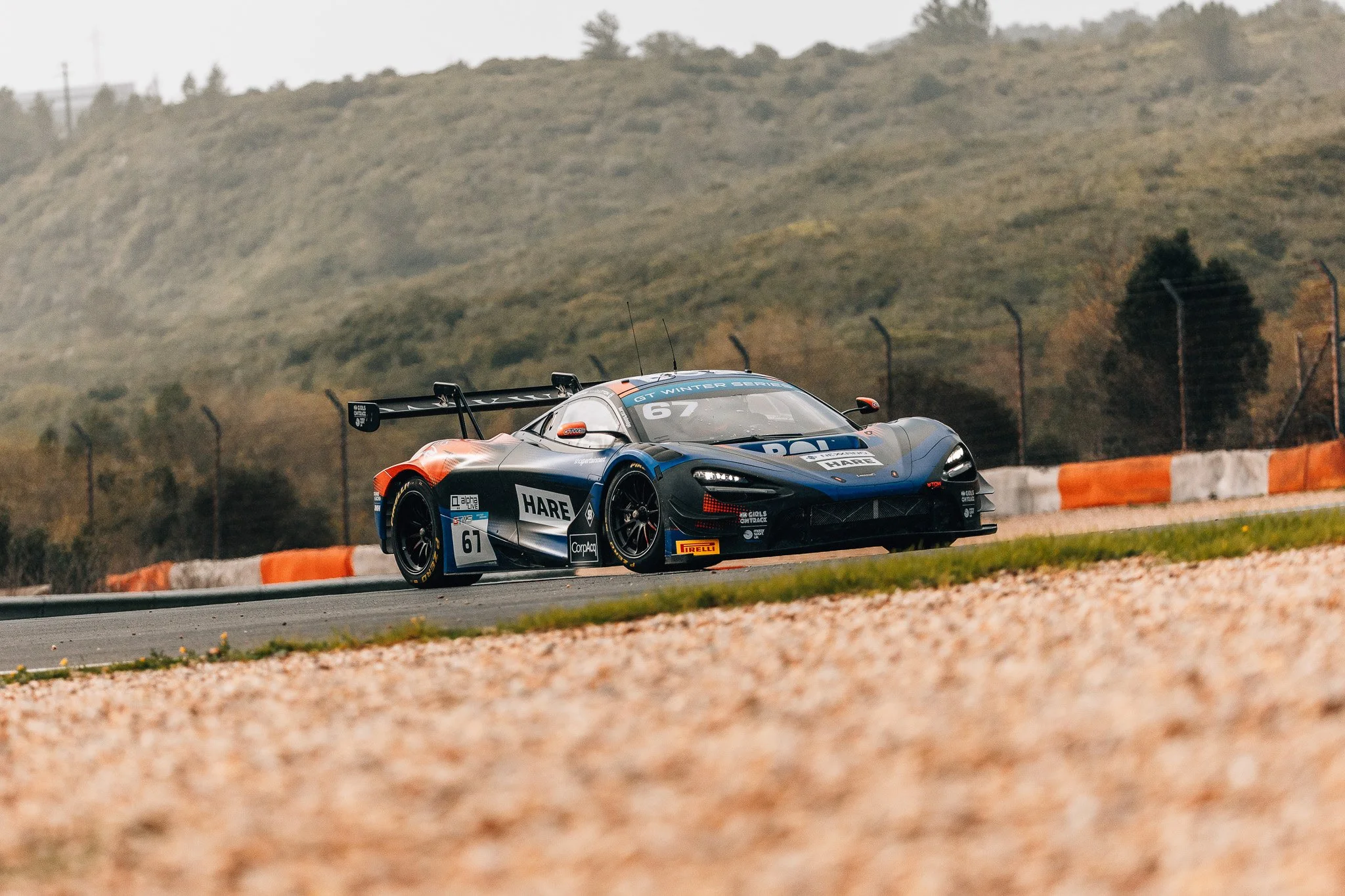 A race car on a racetrack with hills in the background, featuring sponsorship logos and a prominent rear wing.