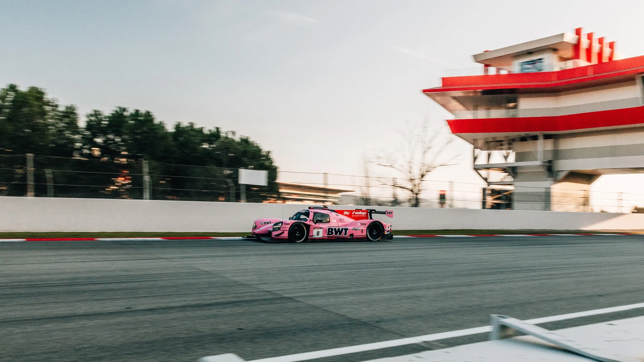 A pink racing car on a race track near a white barrier, with a building and trees in the background during dusk or dawn.
