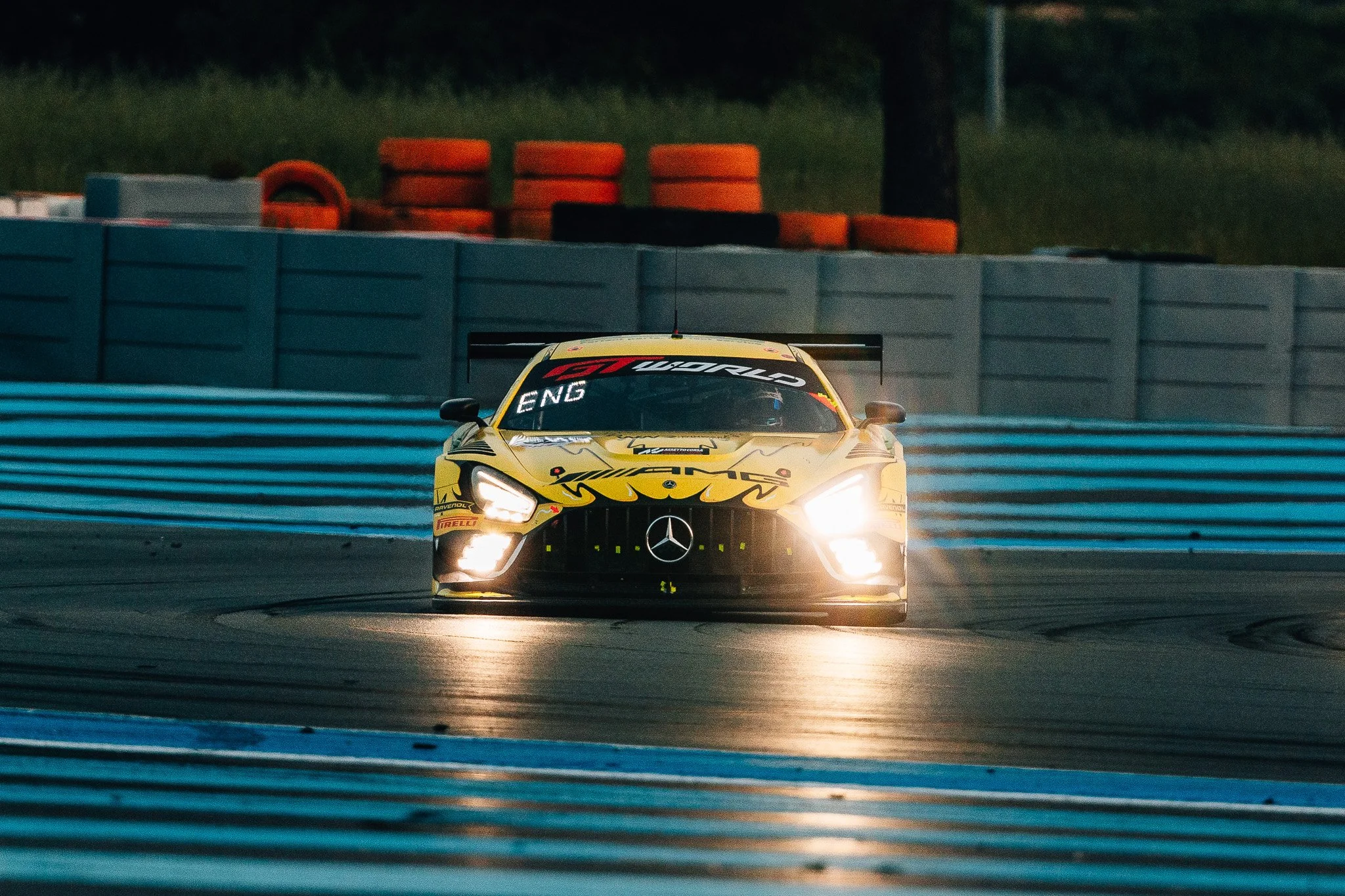 A yellow Mercedes race car speeding on a racetrack at night with headlights on, surrounded by blue safety barriers and orange tire barriers in the background.
